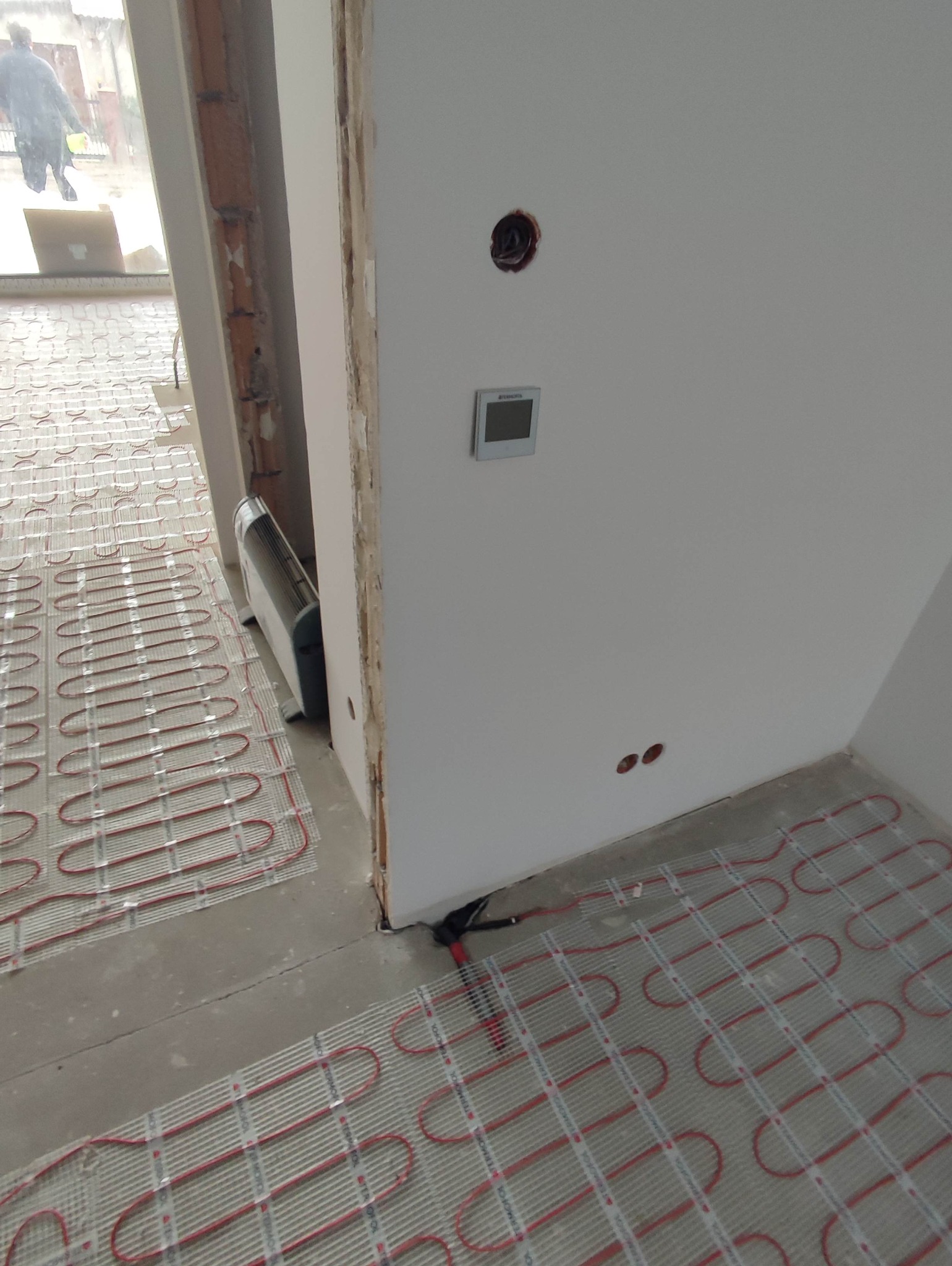 Rozłożona mata grzewcza na podłodze, przygotowana do zalania wylewką. Widoczne kable i regulator temperatury na ścianie. Etap prac instalacyjnych ogrzewania podłogowego.