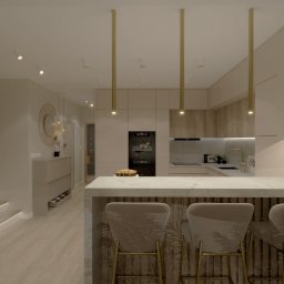 Design Atelier by Maria Słodowska - Elegancka aranżacja wnętrza kuchni z wyspą i hokerami, oświetlona wiszącymi lampami. Widoczna zabudowa kuchenna w jasnych odcieniach i dekoracyjne elementy.