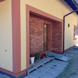 Efekt - Wykończona elewacja domu z ceglaną wstawką przy wejściu i dwukolorową fasadą w odcieniach beżu i brązu, widok z boku, na pierwszym planie fragment chodnika.