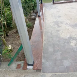 Wykończony taras z kostki brukowej i elementami z czerwonego granitu, widoczne metalowe słupki konstrukcyjne. Detal wykończenia ogrodu.