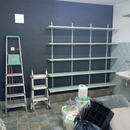 Efekt - Pomieszczenie po malowaniu ścian na ciemny kolor, widoczne drabiny, puszki z farbą i zamontowane białe półki.