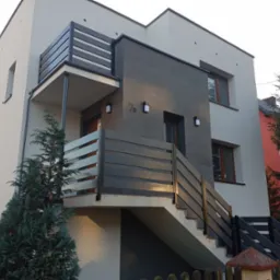 Nowoczesny dom z elewacją w odcieniach szarości i beżu, z drewnianymi balustradami i oświetleniem zewnętrznym. Widok z poziomu gruntu na budynek.