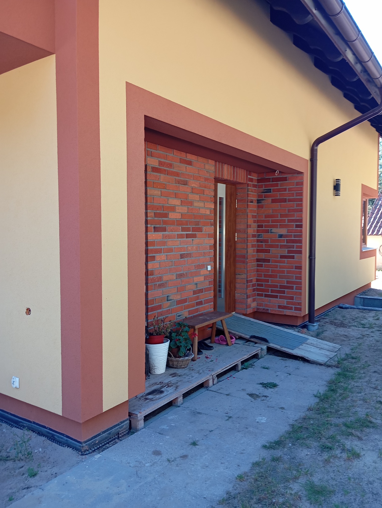 Wykończona elewacja domu z ceglaną wstawką przy wejściu i dwukolorową fasadą w odcieniach beżu i brązu, widok z boku, na pierwszym planie fragment chodnika.