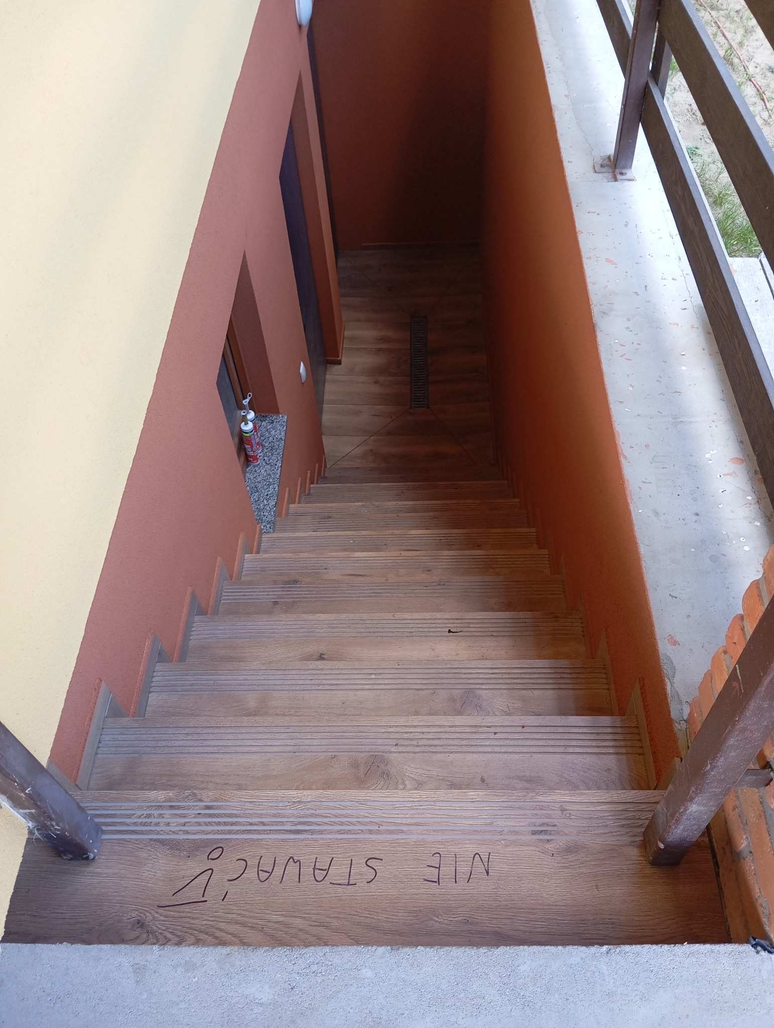 Drewniane schody z napisem 'NIE STAWAĆ!' prowadzą w dół, ściany w odcieniach beżu i brązu. Widok z góry na wykończone wnętrze.