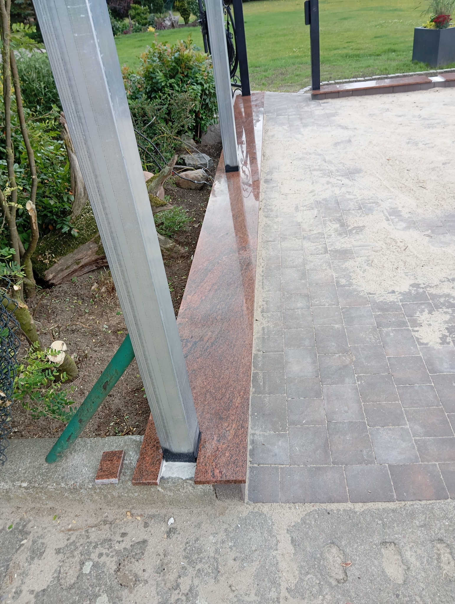 Wykończony taras z kostki brukowej i elementami z czerwonego granitu, widoczne metalowe słupki konstrukcyjne. Detal wykończenia ogrodu.