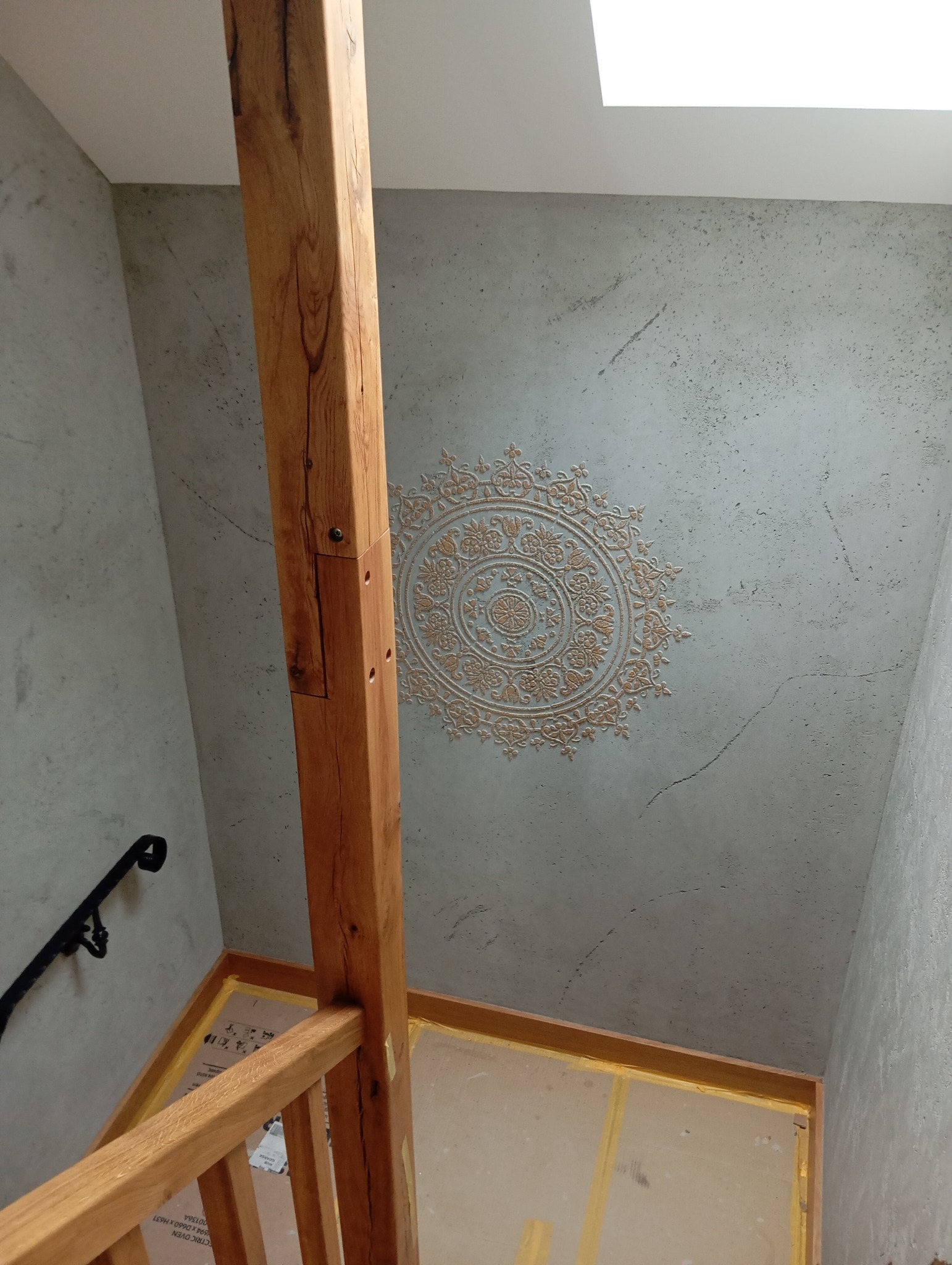 Ściana z dekoracyjnym, okrągłym wzorem w odcieniach beżu, widoczna z perspektywy schodów z drewnianą barierką. Widoczne okno dachowe.