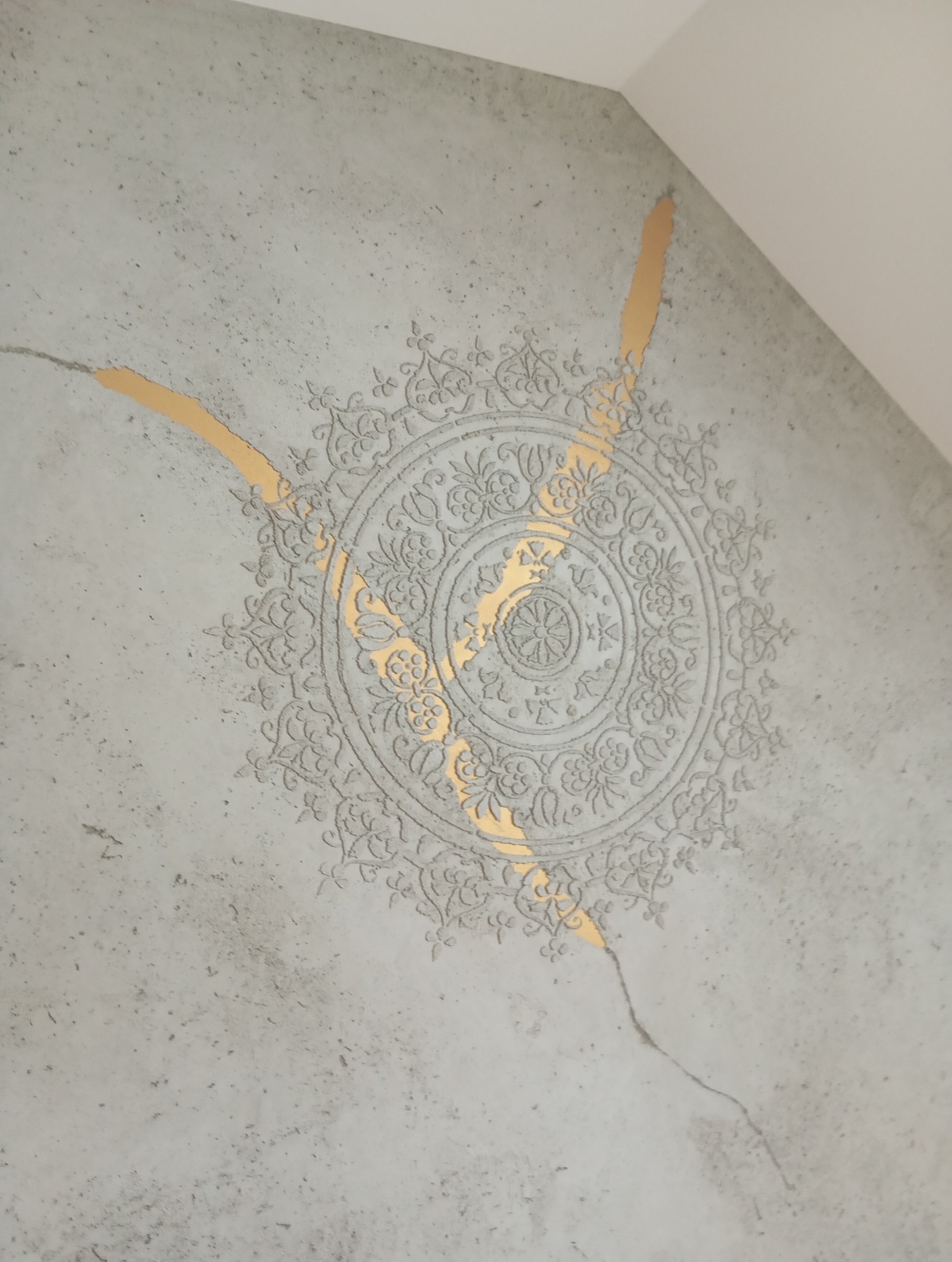 Szara ściana z dekoracyjnym, wypukłym wzorem mandali i złotymi akcentami w szczelinach, tworzącymi efekt kintsugi. Wzór blisko sufitu, widoczna faktura betonu.