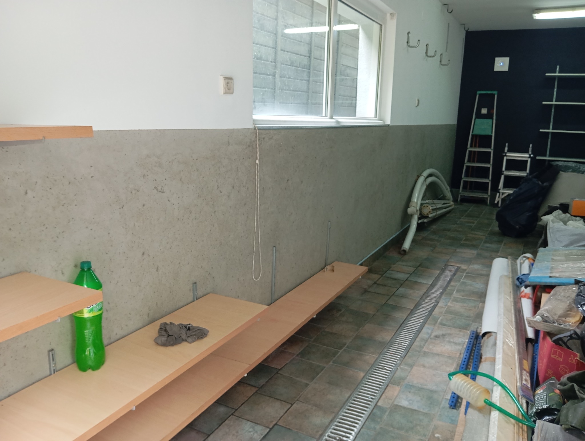 Wnętrze w trakcie prac wykończeniowych: surowa ściana, niedokończone półki, drabina i materiały budowlane. Widoczne elementy instalacji i narzędzia.