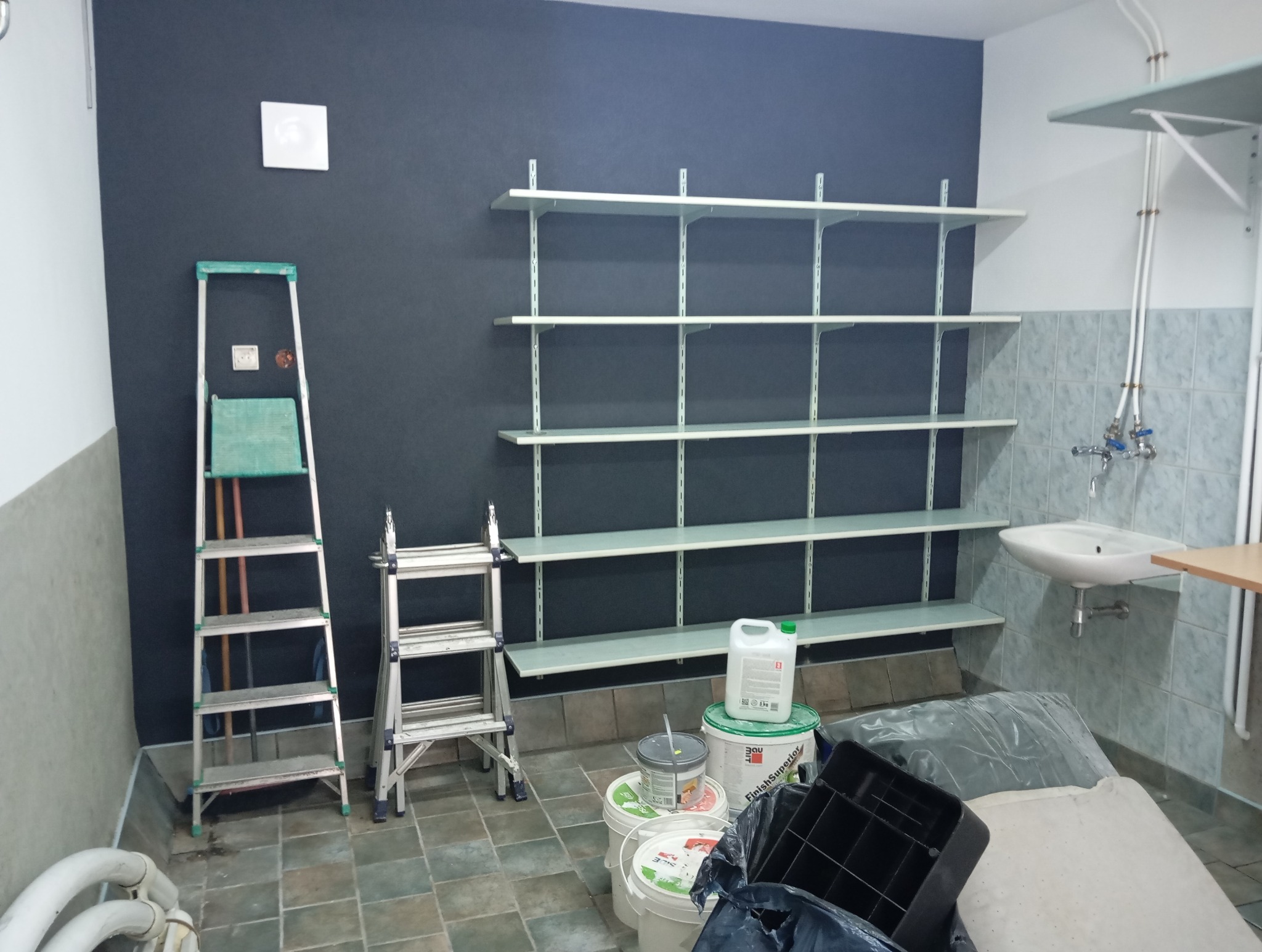 Pomieszczenie po malowaniu ścian na ciemny kolor, widoczne drabiny, puszki z farbą i zamontowane białe półki.