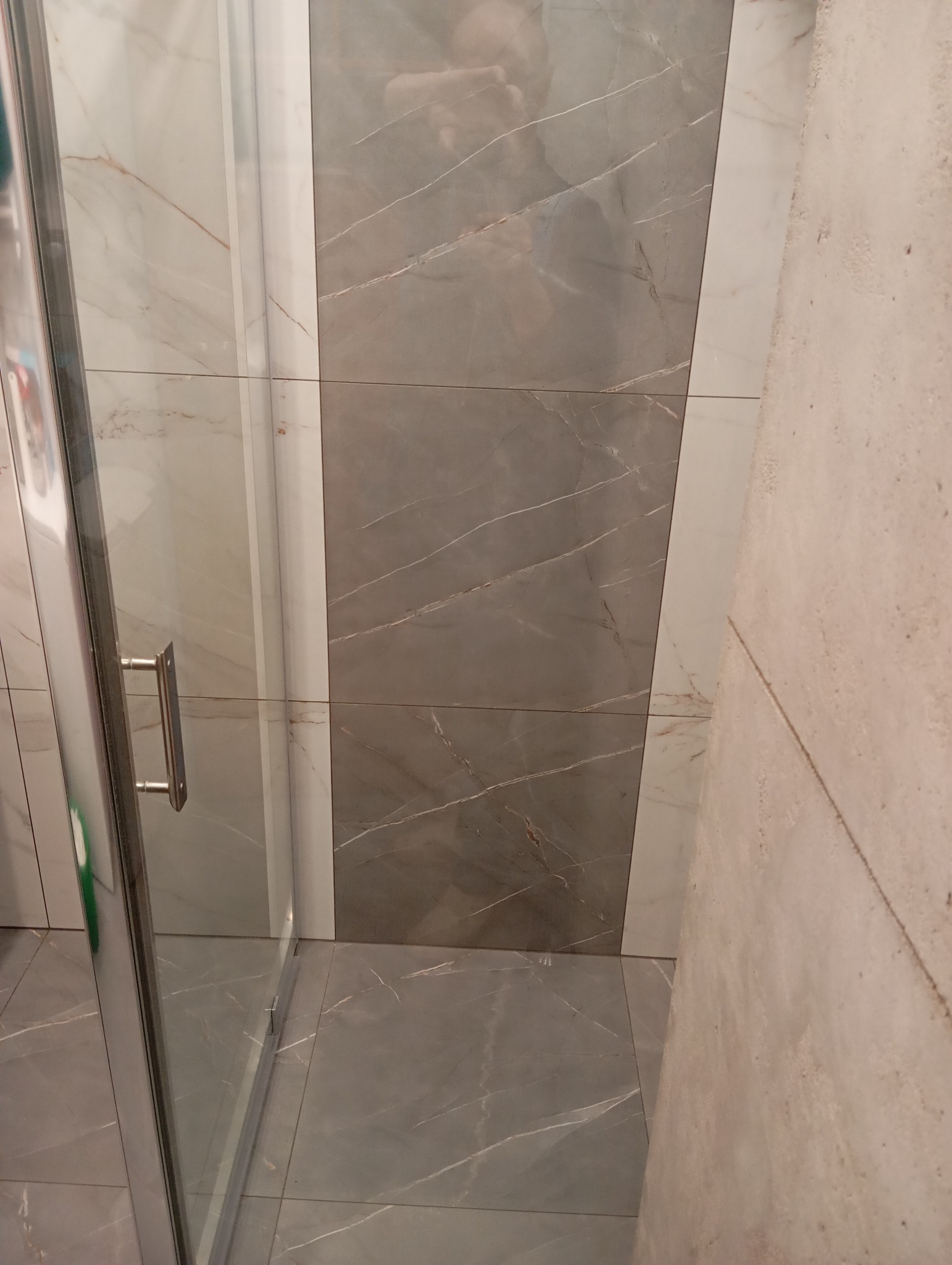 Wykończona łazienka z szarymi i białymi płytkami imitującymi marmur. Widoczny fragment kabiny prysznicowej i surowa ściana.