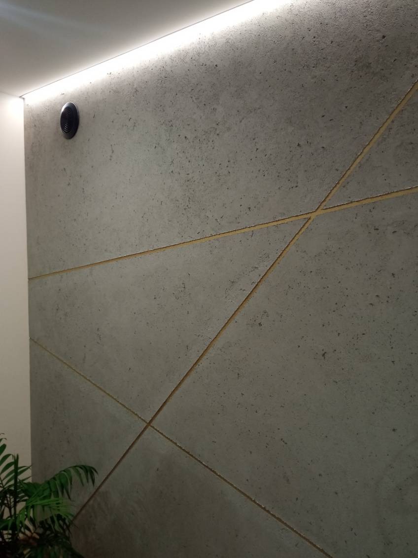 Betonowa ściana z geometrycznym wzorem, oświetlenie LED u góry, fragment rośliny w rogu. Minimalistyczny design, szara kolorystyka, czarny punkt oświetleniowy.