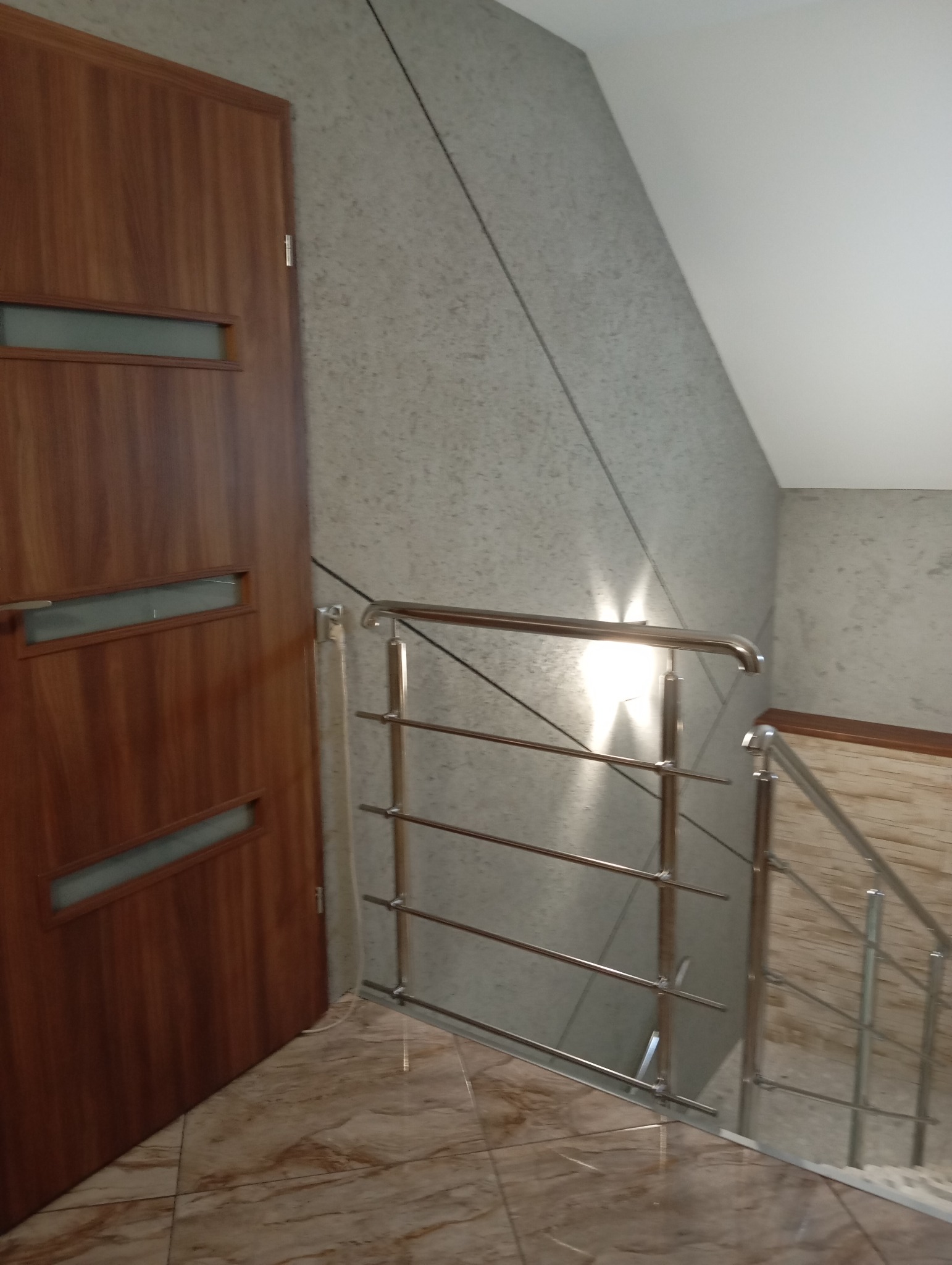 Klatka schodowa po remoncie: brązowe drzwi z przeszkleniami, szara ściana z fakturą, metalowa barierka schodów i marmurowe płytki podłogowe.