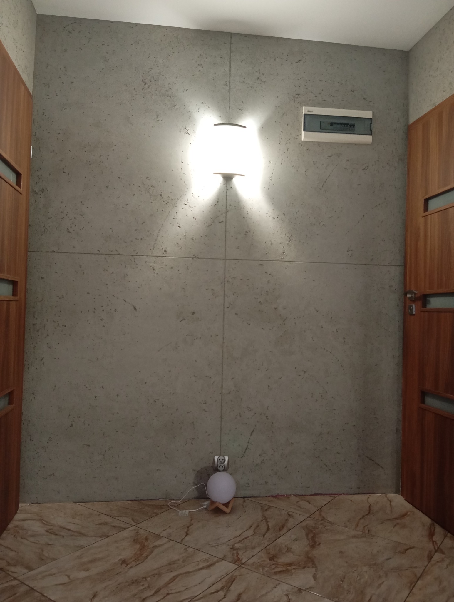 Ściana z efektem betonu podzielona na panele, oświetlona kinkietem. Na podłodze brązowe płytki imitujące marmur. Widoczna lampka nocna i skrzynka elektryczna.