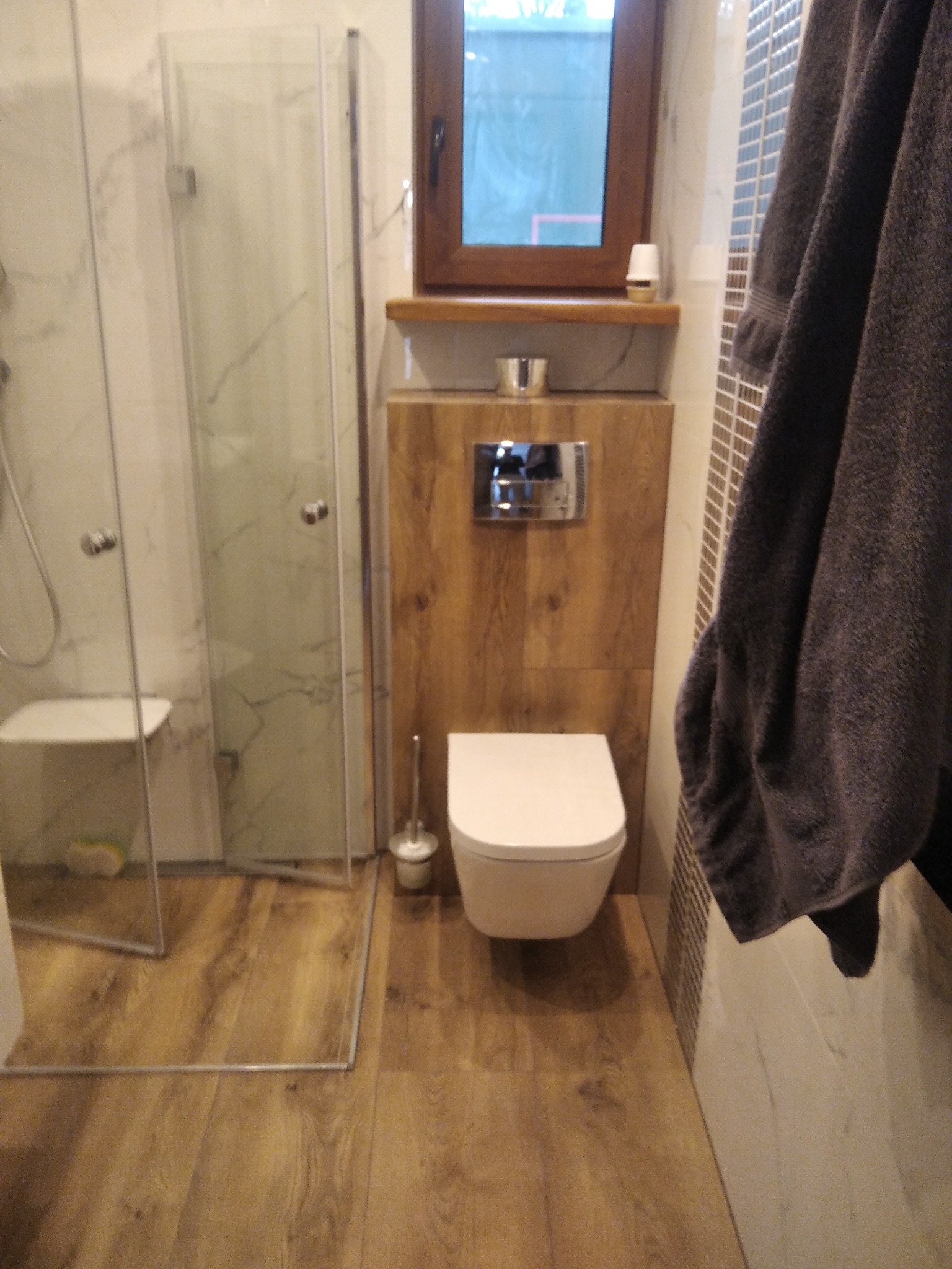 Nowoczesna łazienka z kabiną prysznicową, drewnianą podłogą i toaletą podwieszaną. Szary ręcznik dodaje przytulności.