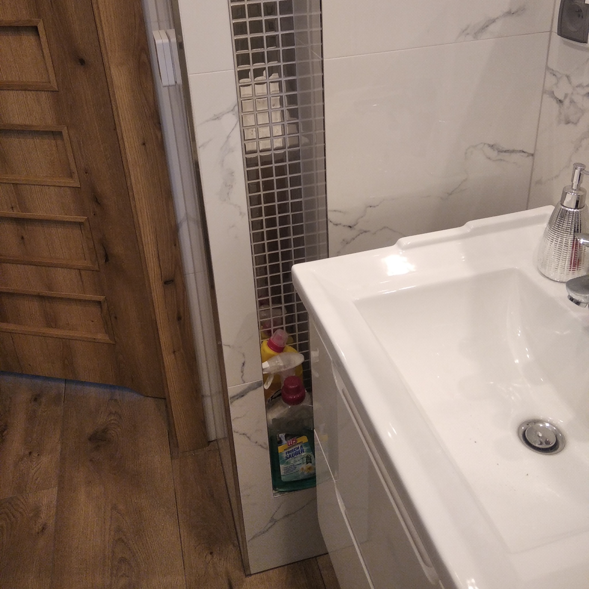 Biała umywalka nablatowa z baterią i dozownikiem mydła, obok drewniane drzwi i ściana wyłożona płytkami imitującymi marmur z mozaikowym pasem.