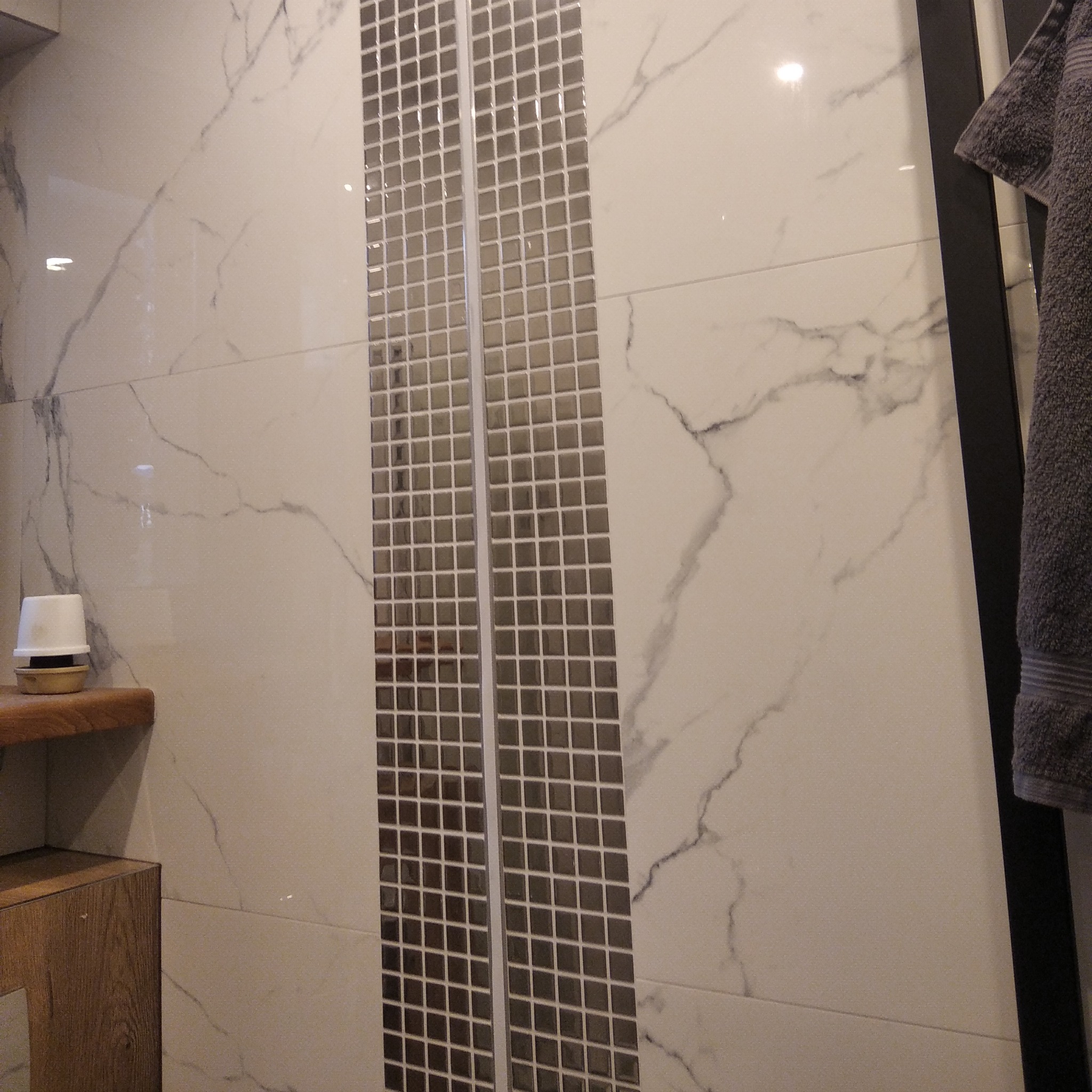 Wykończona ściana łazienki z białymi płytkami imitującymi marmur i dwoma pionowymi pasami mozaiki. Widoczny fragment szafki pod umywalkę i ręcznik.