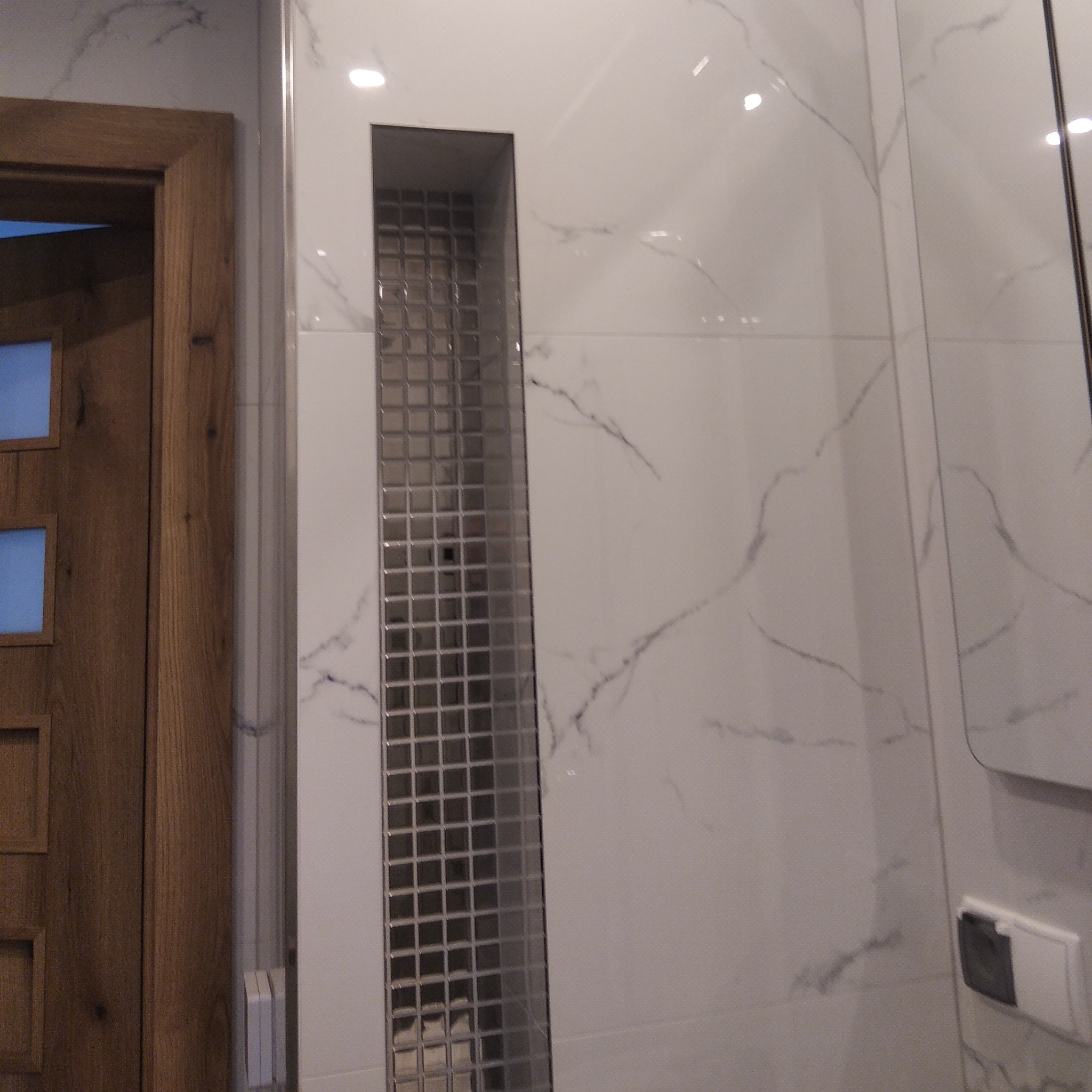 Biała ściana z płytek imitujących marmur z wnęką wyłożoną mozaiką, obok drewniane drzwi. Minimalistyczne wykończenie łazienki.