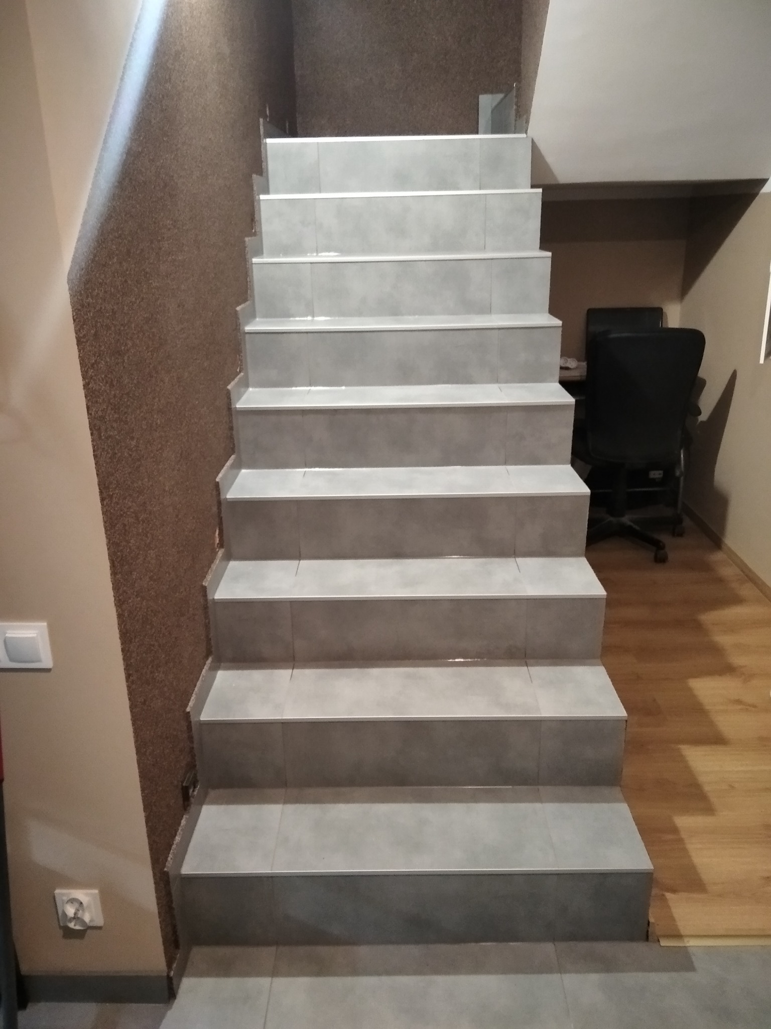 Wykończone schody wewnętrzne z szarymi płytkami, widoczne z dołu. W tle ściana z dekoracyjnym tynkiem i fragment biura z krzesłem.