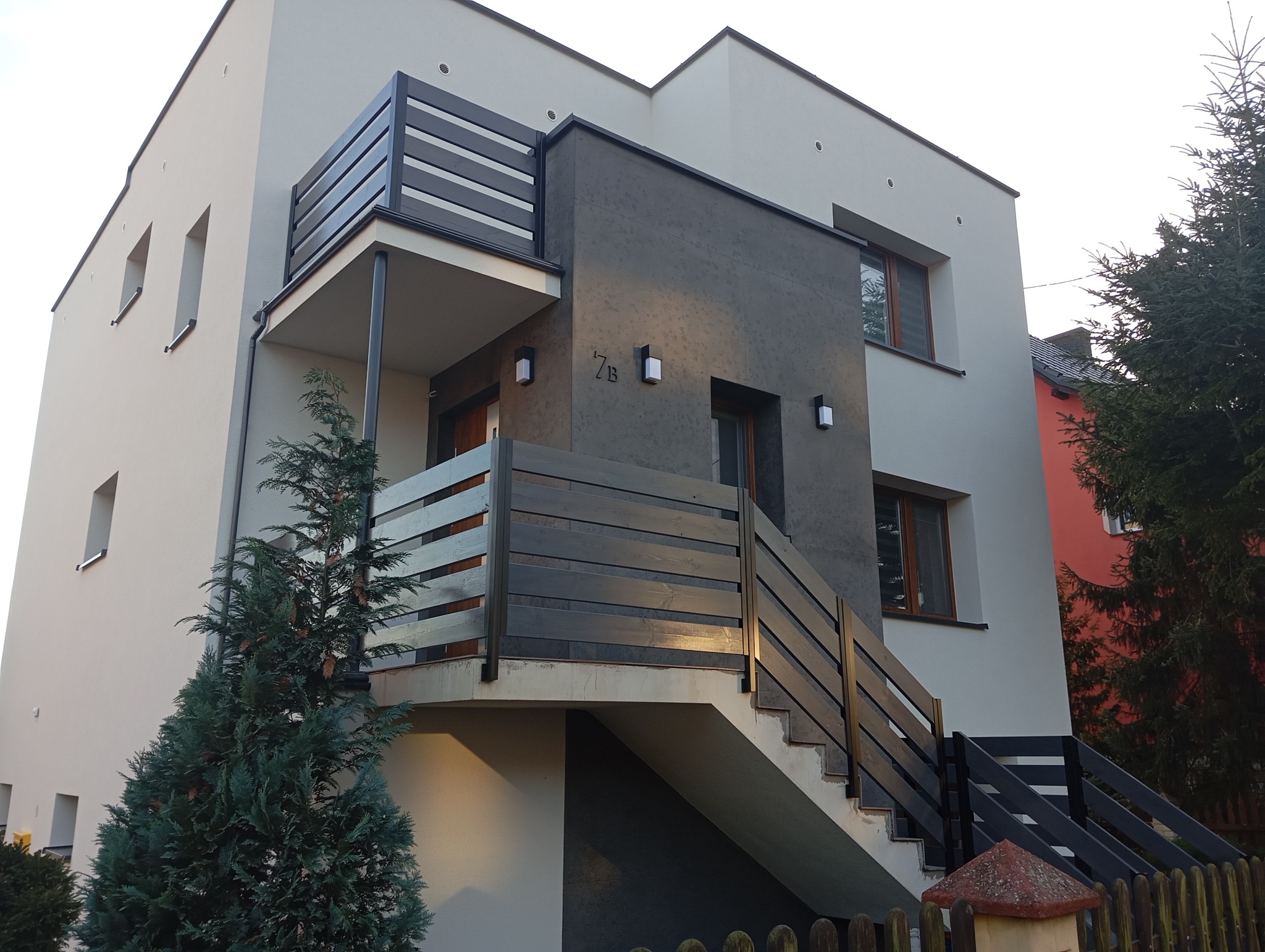 Nowoczesny dom z elewacją w odcieniach szarości i beżu, z drewnianymi balustradami i oświetleniem zewnętrznym. Widok z poziomu gruntu na budynek.