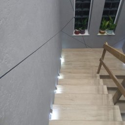 REMONTY PERFEKTO - Schody z drewnianymi stopniami i podświetleniem LED wzdłuż ściany. Szara ściana z dekoracyjnymi liniami. Dwa okna z roślinami na parapecie.