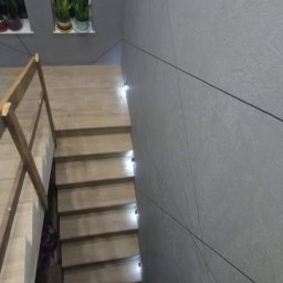 REMONTY PERFEKTO - Schody drewniane z podświetleniem LED w szarej klatce schodowej z geometrycznym wzorem na ścianie i roślinami w oknach. Ujęcie z góry.