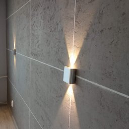 REMONTY PERFEKTO - Dwa nowoczesne, srebrne kinkiety ścienne, emitujące stożkowe światło na szarej ścianie z fakturą betonu, podkreślone białymi listwami. Widok z perspektywy.