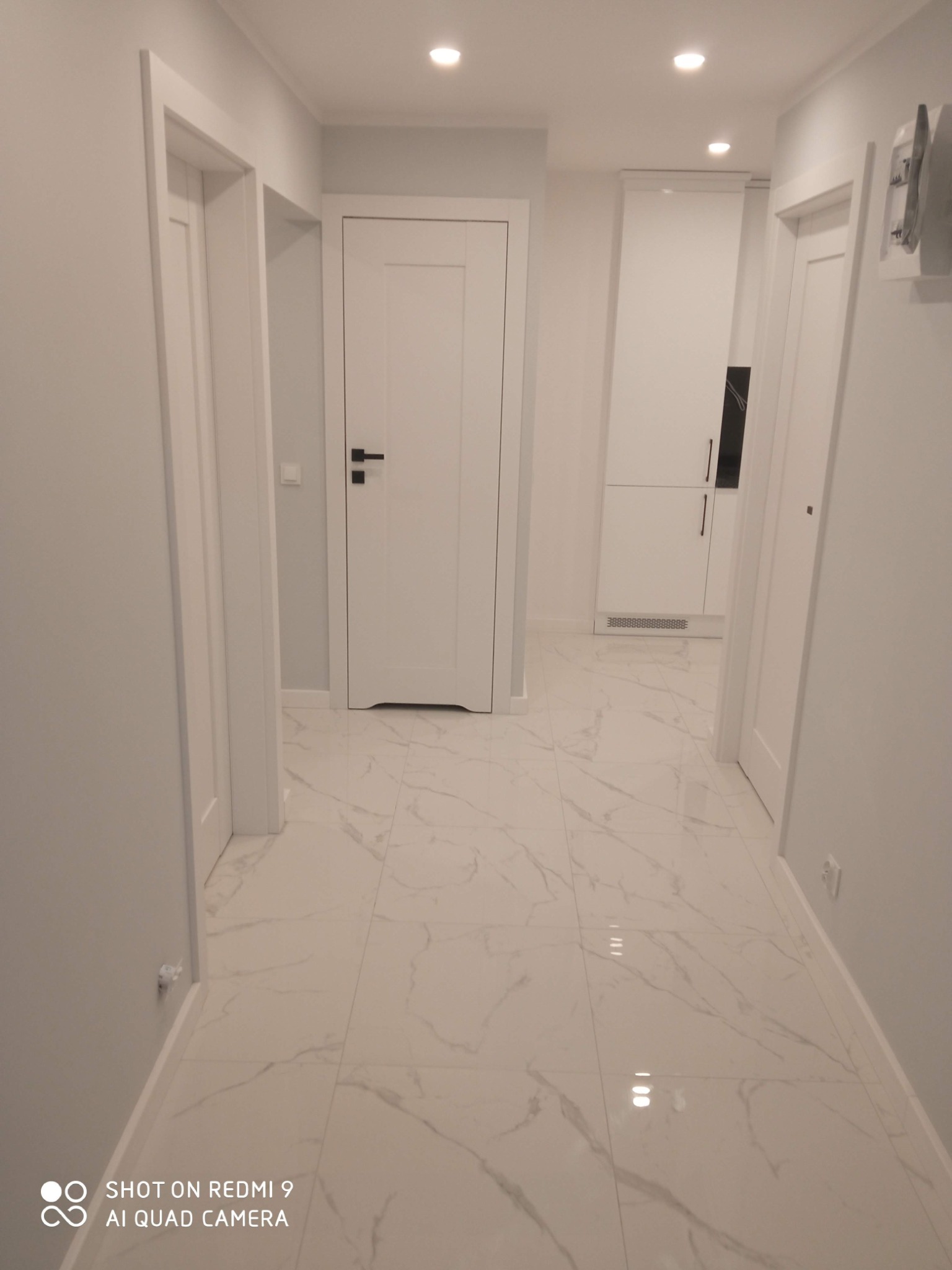 Jasne wnętrze korytarza z białymi drzwiami, marmurową podłogą i punktowym oświetleniem. Minimalistyczny design z akcentami czerni w postaci klamek.