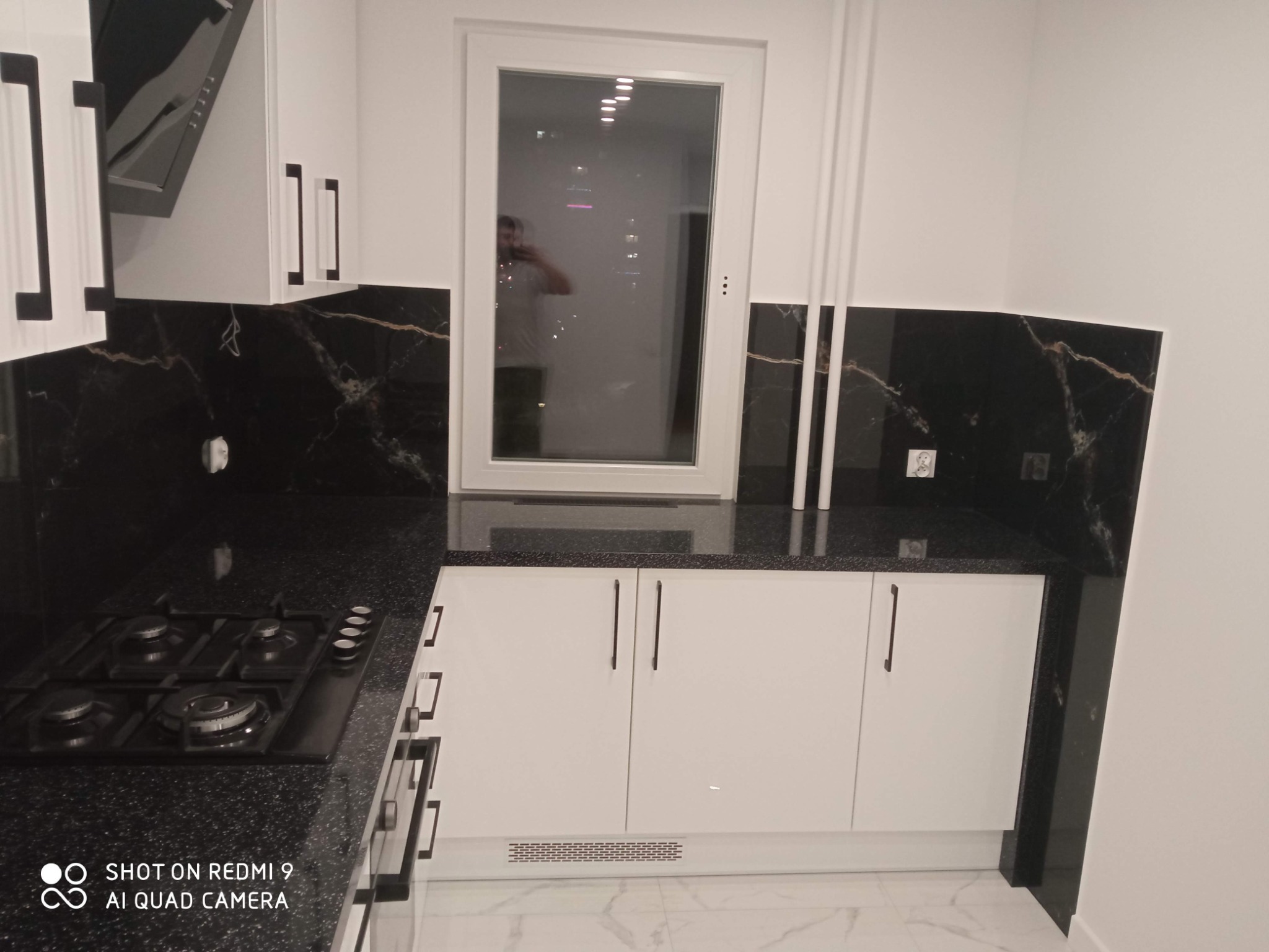 Nowoczesna kuchnia z białymi szafkami, czarnym blatem imitującym marmur i płytą gazową. W oknie odbija się osoba robiąca zdjęcie.