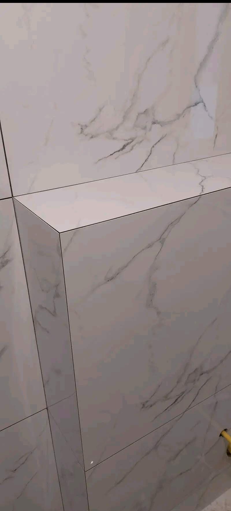 Wykończona ściana i półka w łazience, pokryte białymi płytkami imitującymi marmur. Widoczne fugi i narożnik półki. Minimalistyczny design.