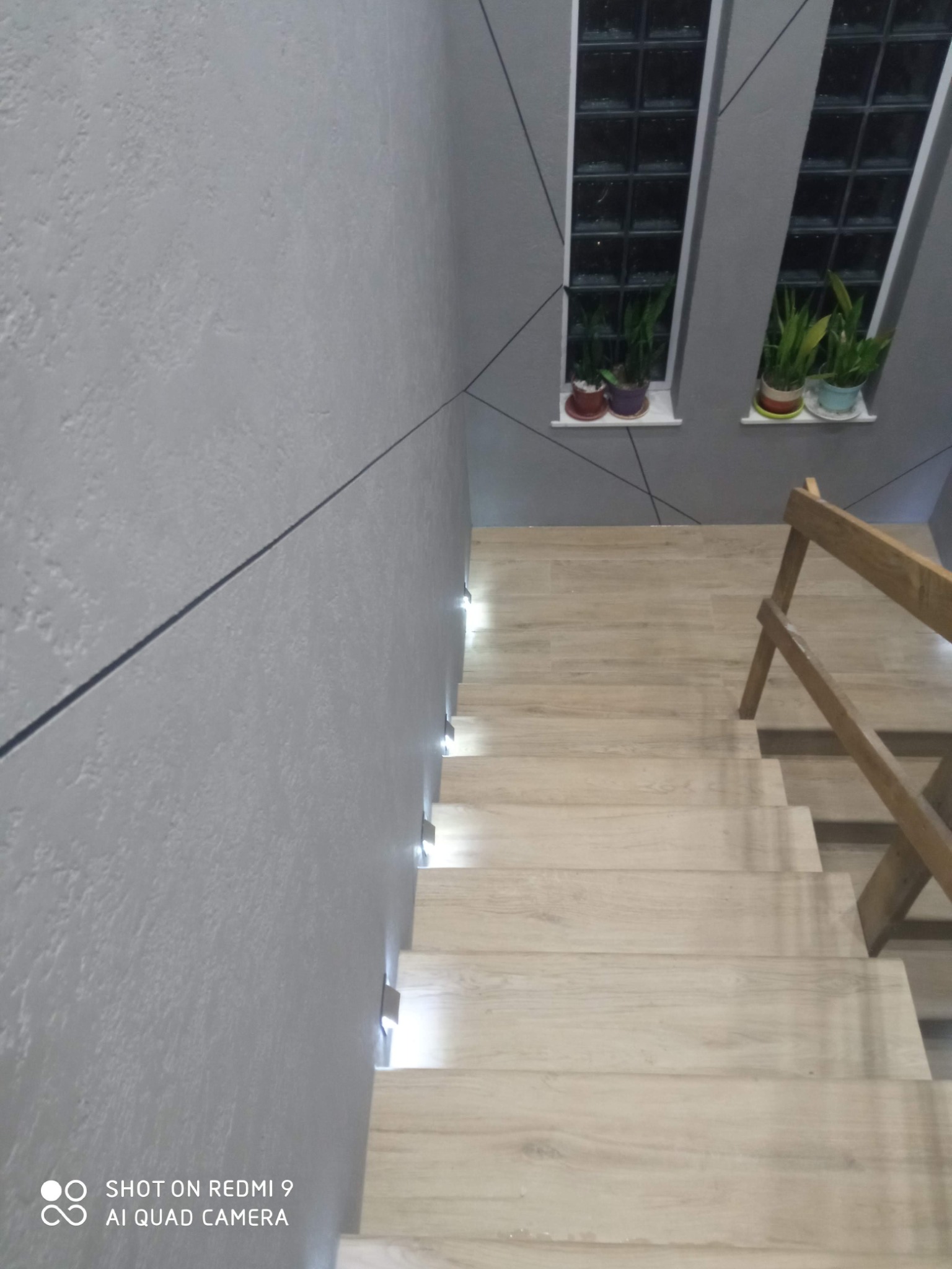 Schody z drewnianymi stopniami i podświetleniem LED wzdłuż ściany. Szara ściana z dekoracyjnymi liniami. Dwa okna z roślinami na parapecie.