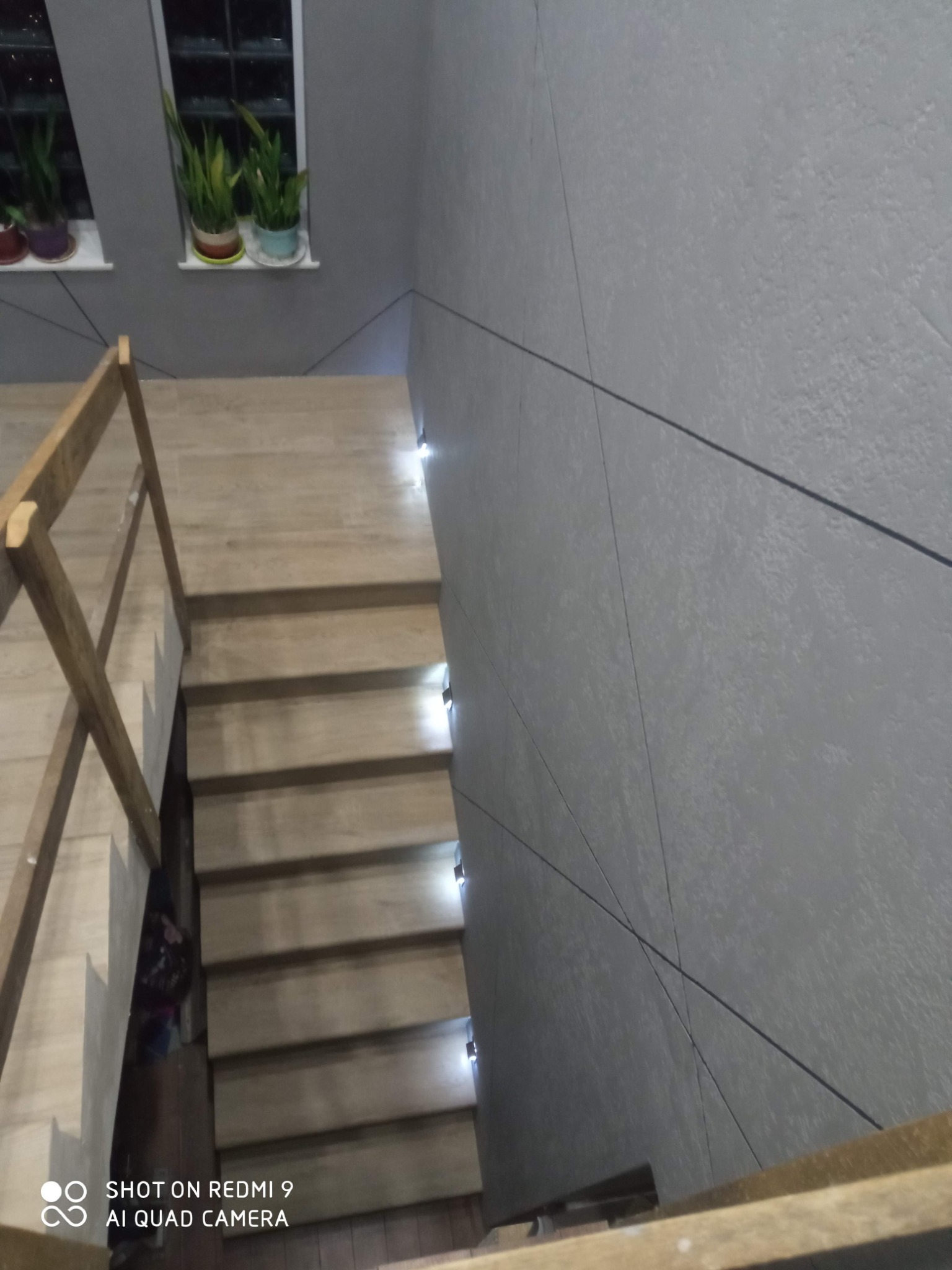 Schody drewniane z podświetleniem LED w szarej klatce schodowej z geometrycznym wzorem na ścianie i roślinami w oknach. Ujęcie z góry.