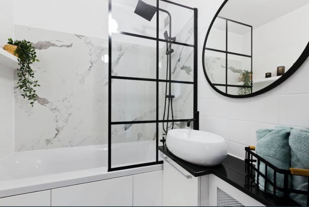 Nowoczesna łazienka z wanną i kabiną prysznicową typu walk-in, białe płytki imitujące marmur, owalne lustro w czarnej ramie i minimalistyczna umywalka.
