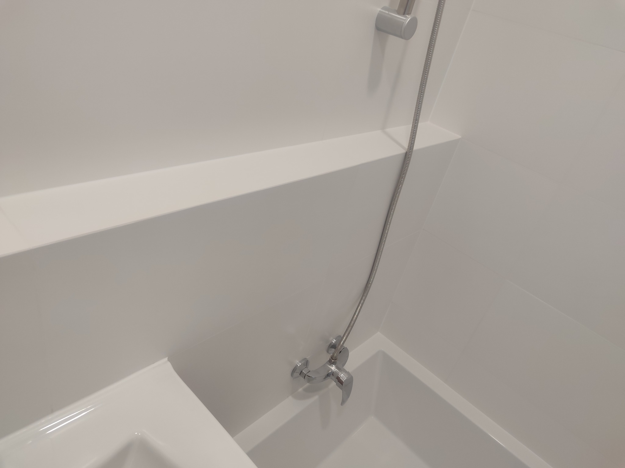 Biała, minimalistyczna łazienka z wanną, chromowaną baterią i półką w zabudowie. Nowoczesne wykończenie, czyste linie i brak dekoracji.