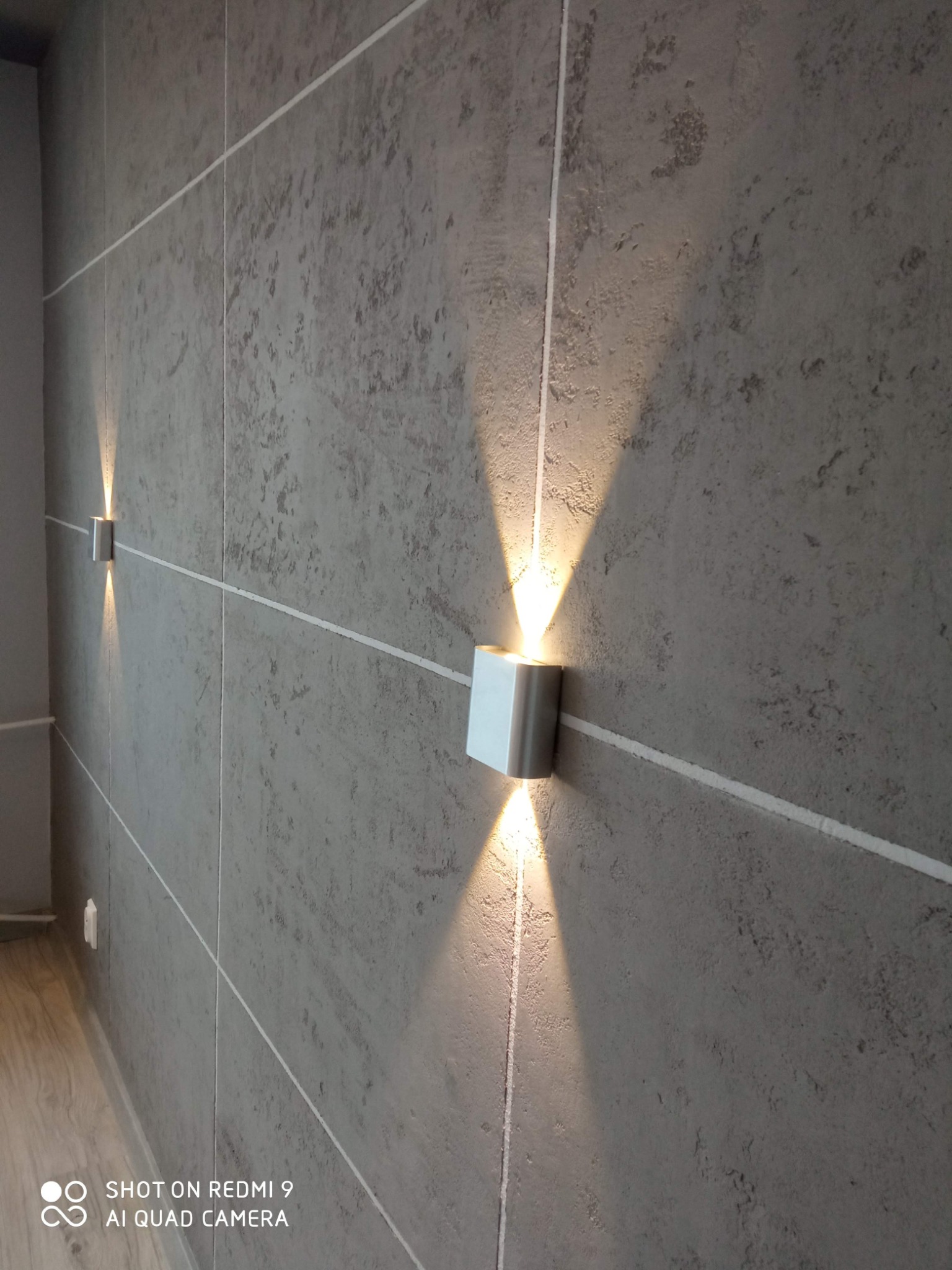 Dwa nowoczesne, srebrne kinkiety ścienne, emitujące stożkowe światło na szarej ścianie z fakturą betonu, podkreślone białymi listwami. Widok z perspektywy.