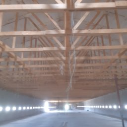 DAMIAN DYMEK EKO DOM IZOLACJE NATRYSKOWE - Konstrukcja drewniana dachu od wewnątrz, widoczne krokwie i izolacja, w tle ściana z oknami. Prace budowlane wewnątrz budynku.