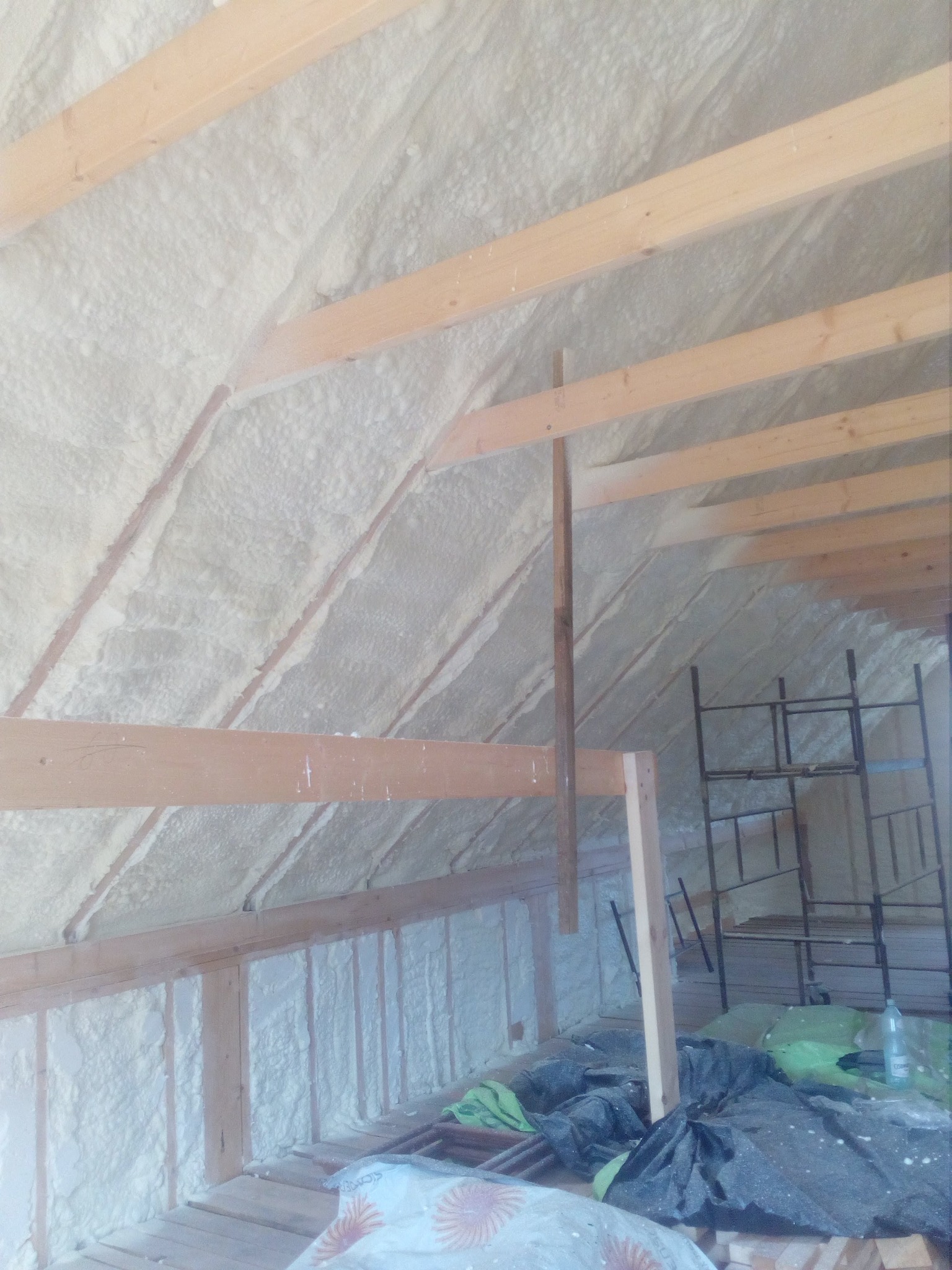 Poddasze z drewnianą konstrukcją i białą pianką izolacyjną, widoczne rusztowanie i materiały budowlane. Ocieplenie pianką PUR w trakcie realizacji.