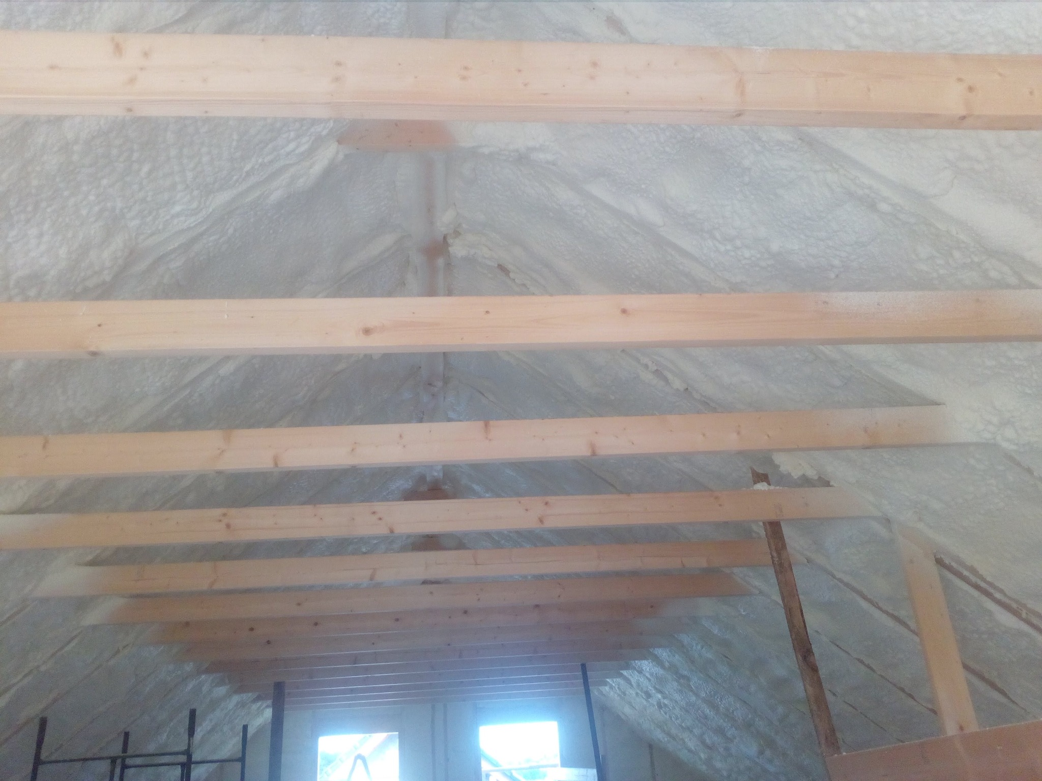 Poddasze z drewnianą konstrukcją i izolacją pianką PUR, widok z dołu. Biała pianka szczelnie wypełnia przestrzeń między krokwiami. W tle okna.