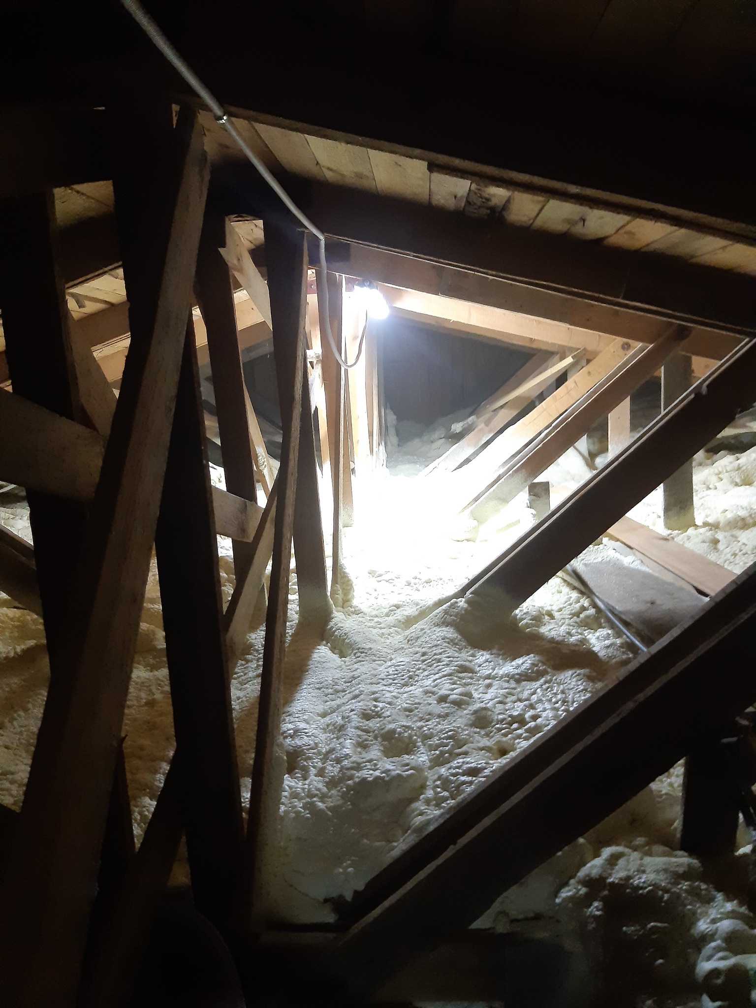 Poddasze z drewnianą konstrukcją wypełnione białą pianką izolacyjną, widok z dołu, oświetlone punktowo. Widoczne elementy instalacji elektrycznej.
