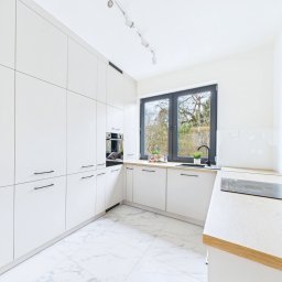 AD-MAR Bartłomiej Żmuda - Biała kuchnia na wymiar z czarnymi akcentami: minimalistyczne fronty, marmurowe płytki podłogowe i okno z widokiem na zieleń. Nowoczesny design.