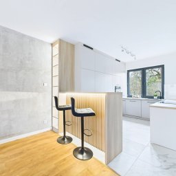 AD-MAR Bartłomiej Żmuda - Nowoczesna, jasna kuchnia z wyspą oświetloną LED, drewnianymi akcentami i dwoma czarnymi hokerami. Minimalistyczny design, okno z widokiem na zieleń.