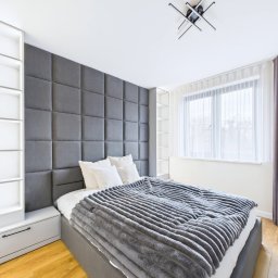AD-MAR Bartłomiej Żmuda - Nowoczesna sypialnia z szarą tapicerowaną ścianą za łóżkiem, białe półki w zabudowie i drewniana podłoga. Minimalistyczny design i stonowana kolorystyka.