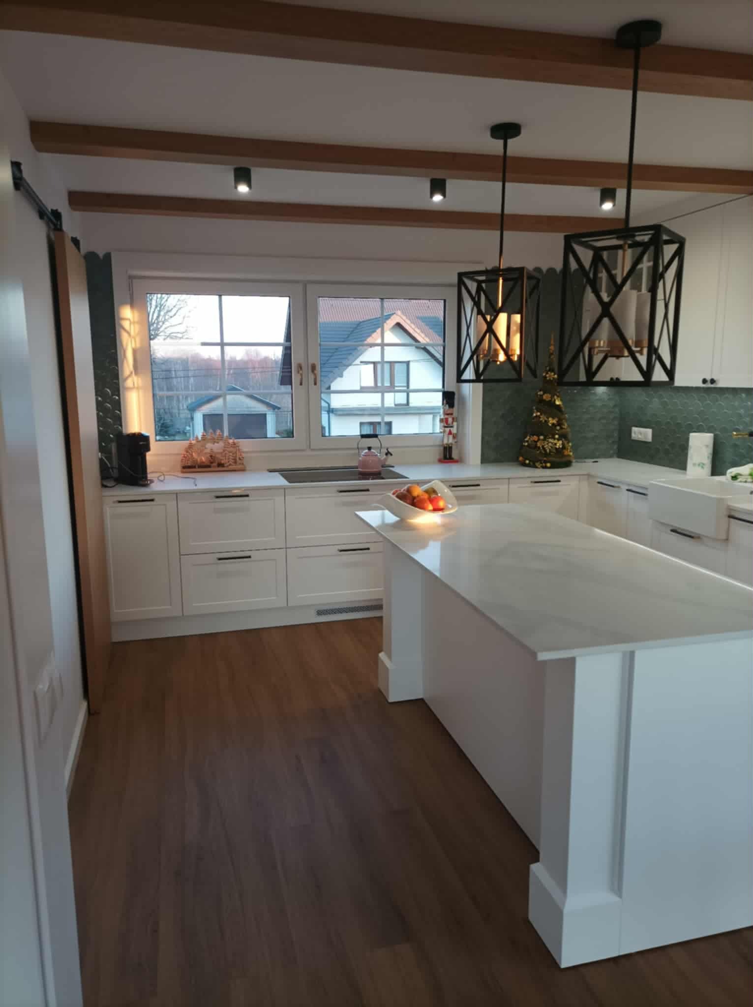 Jasna kuchnia z wyspą i drewnianymi belkami sufitowymi. Dwa wiszące, czarne, geometryczne klosze lamp. Za oknem widoczny dom.