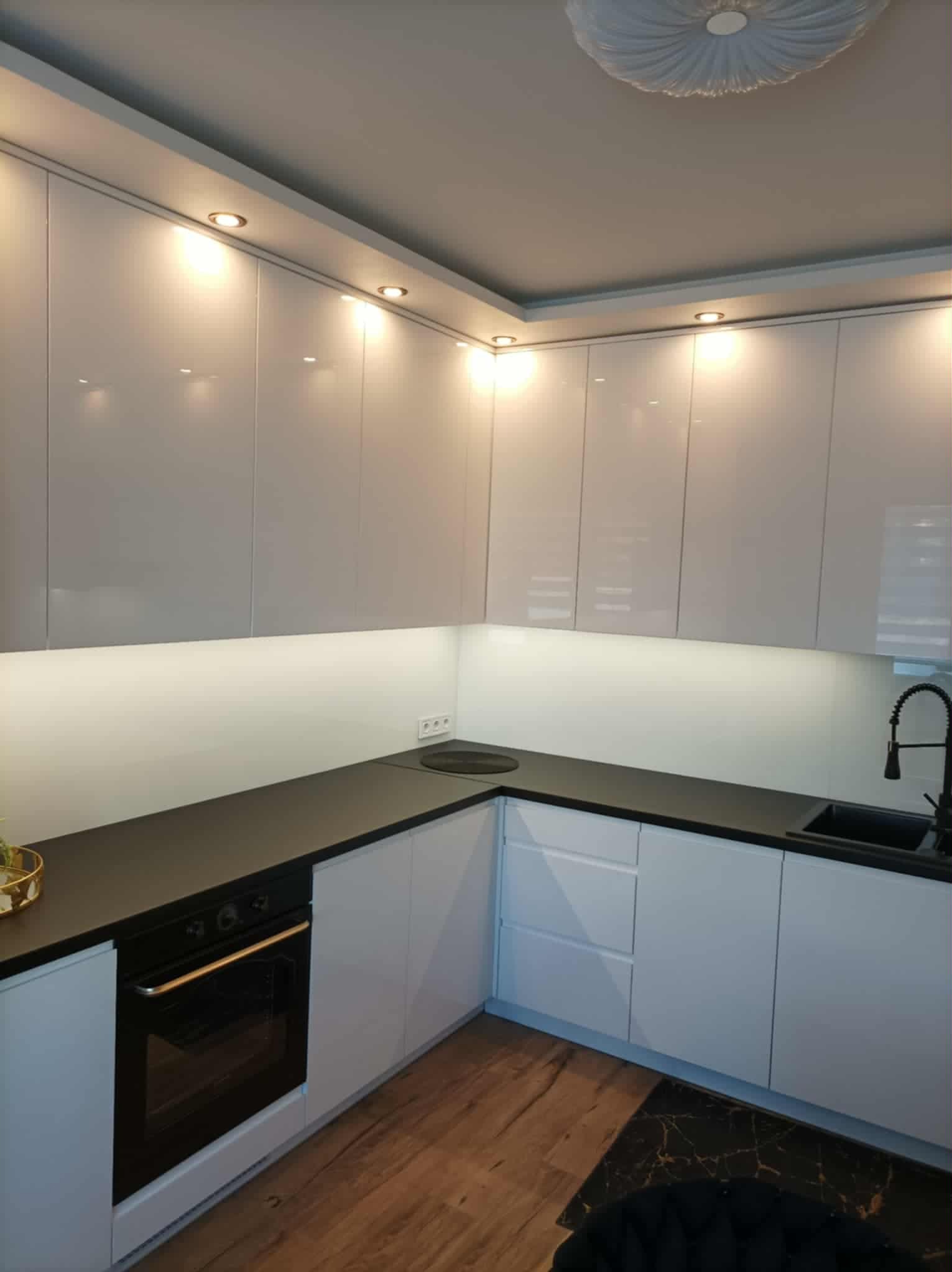Biała, nowoczesna kuchnia z połyskiem, czarnym blatem i oświetleniem LED podszafkowym. Widoczny piekarnik, zlew i płyta grzewcza. Minimalistyczny design.
