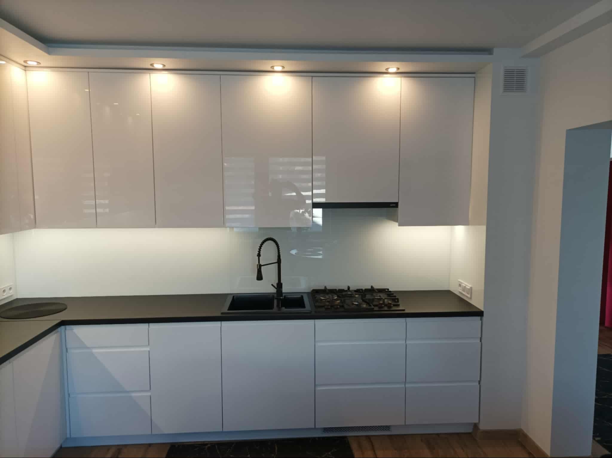 Nowoczesna, biała kuchnia z czarnym blatem i armaturą. Górne szafki z połyskiem, oświetlenie punktowe. Widoczny fragment płyty gazowej i zlewozmywaka.