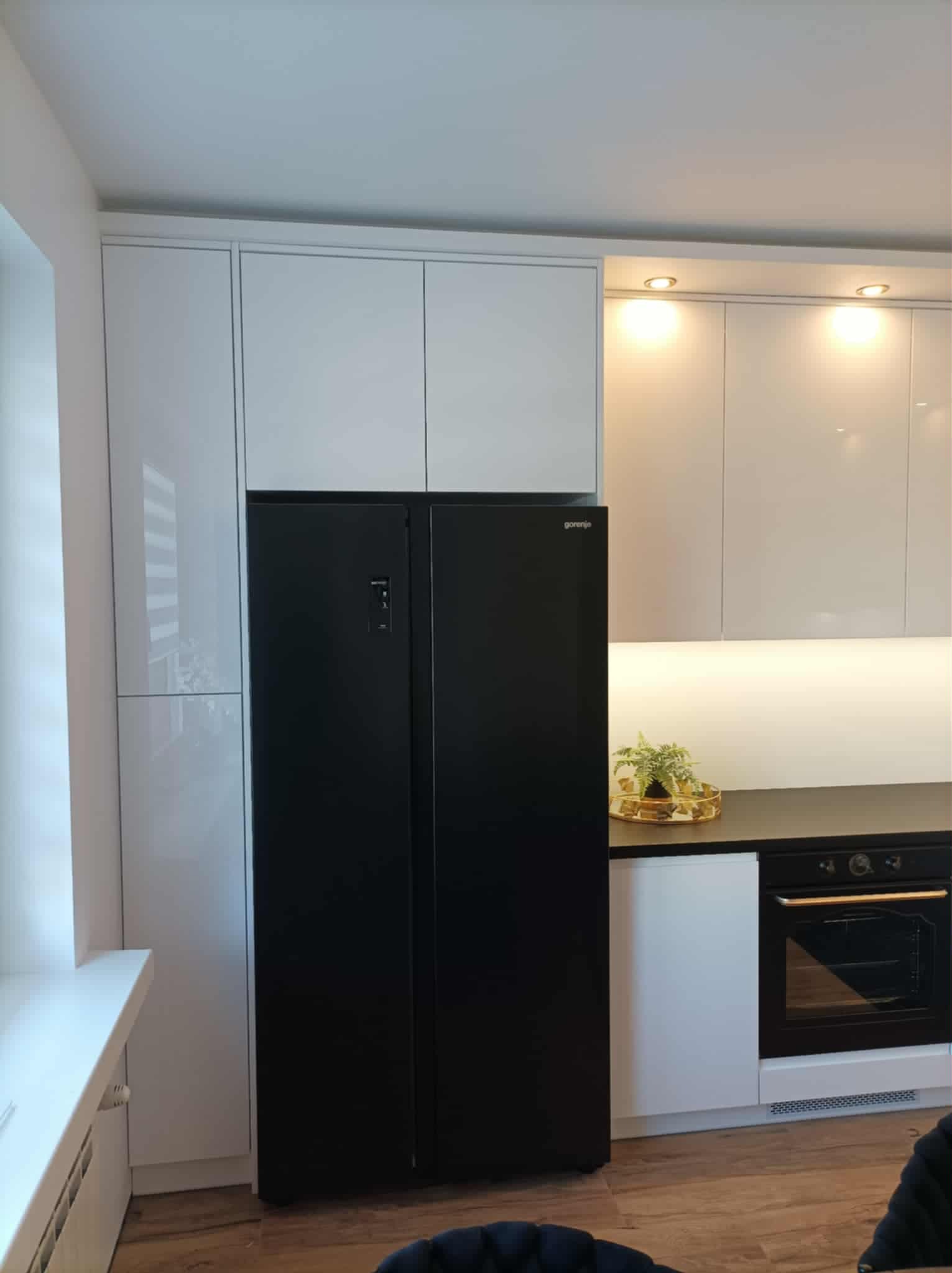 Biała zabudowa kuchenna z czarną lodówką side-by-side Gorenje. Górne szafki podświetlone, blat ciemny, na nim dekoracja. Widoczny piekarnik w zabudowie.
