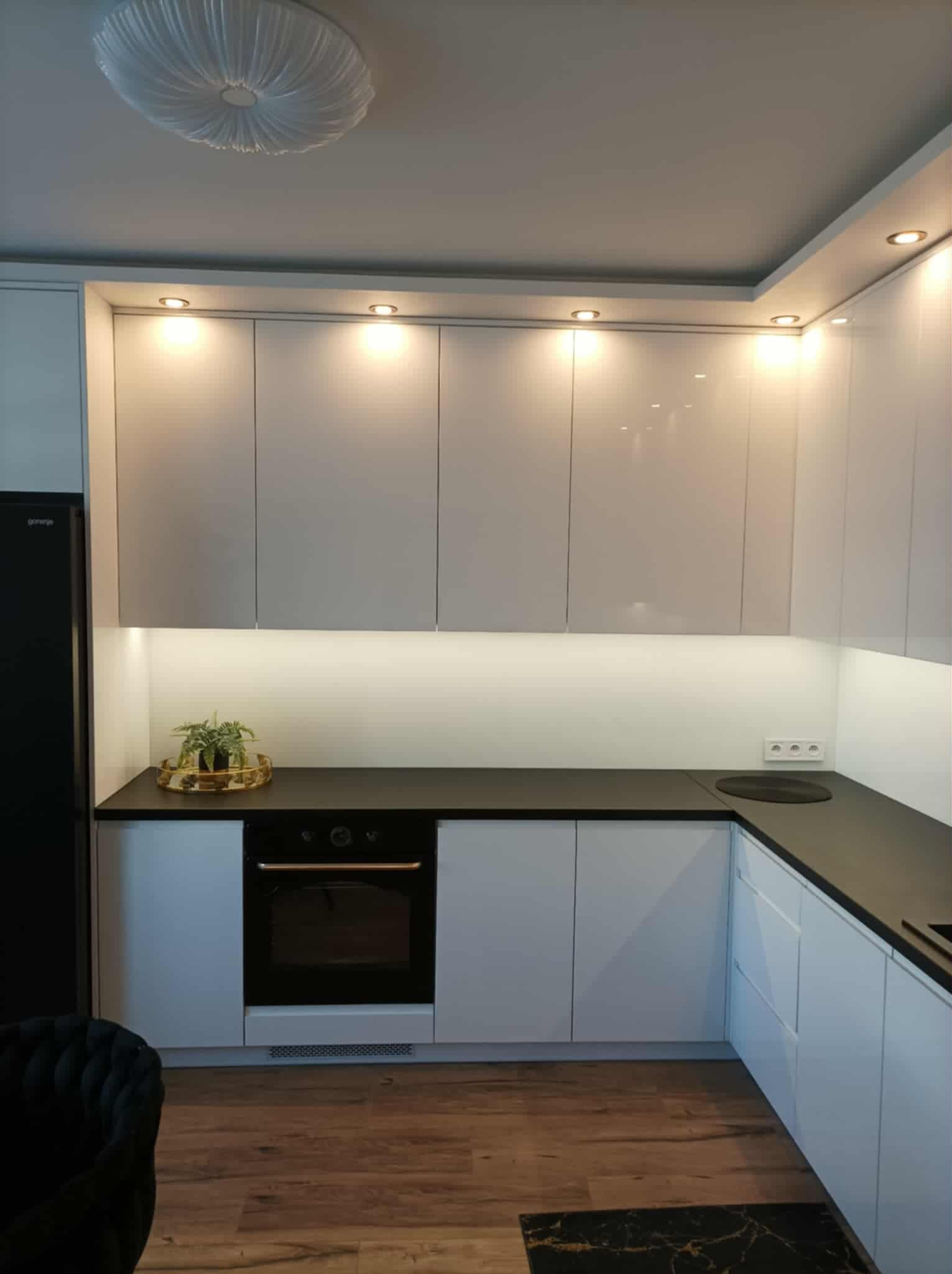 Nowoczesna kuchnia z białymi szafkami, czarnym blatem i oświetleniem LED. Na blacie roślina w złotej doniczce. Widoczny fragment czarnego fotela.