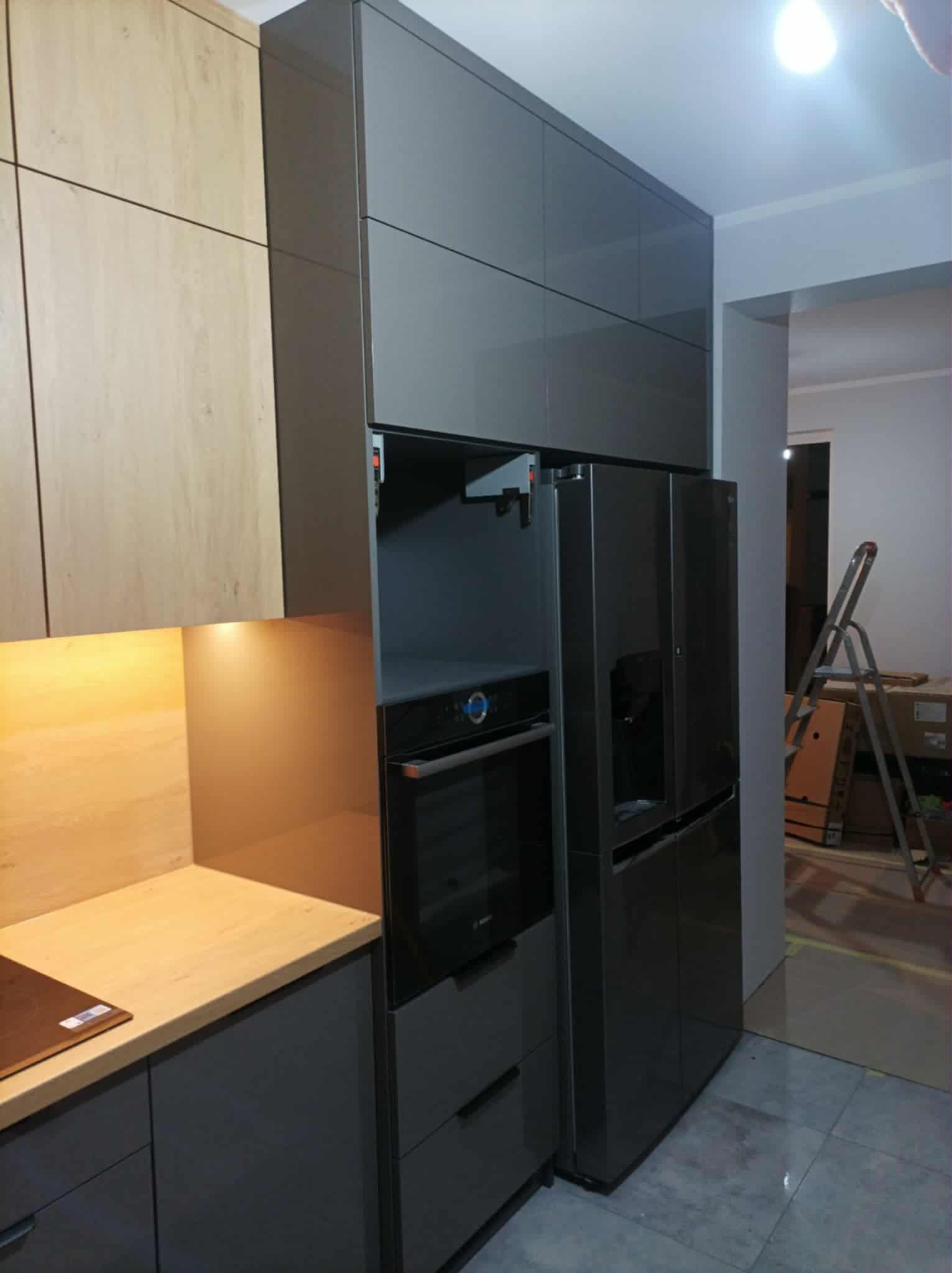 Szara zabudowa kuchenna z piekarnikiem i lodówką side-by-side, jasne drewniane szafki górne, widoczna drabina w tle, nowoczesny design.