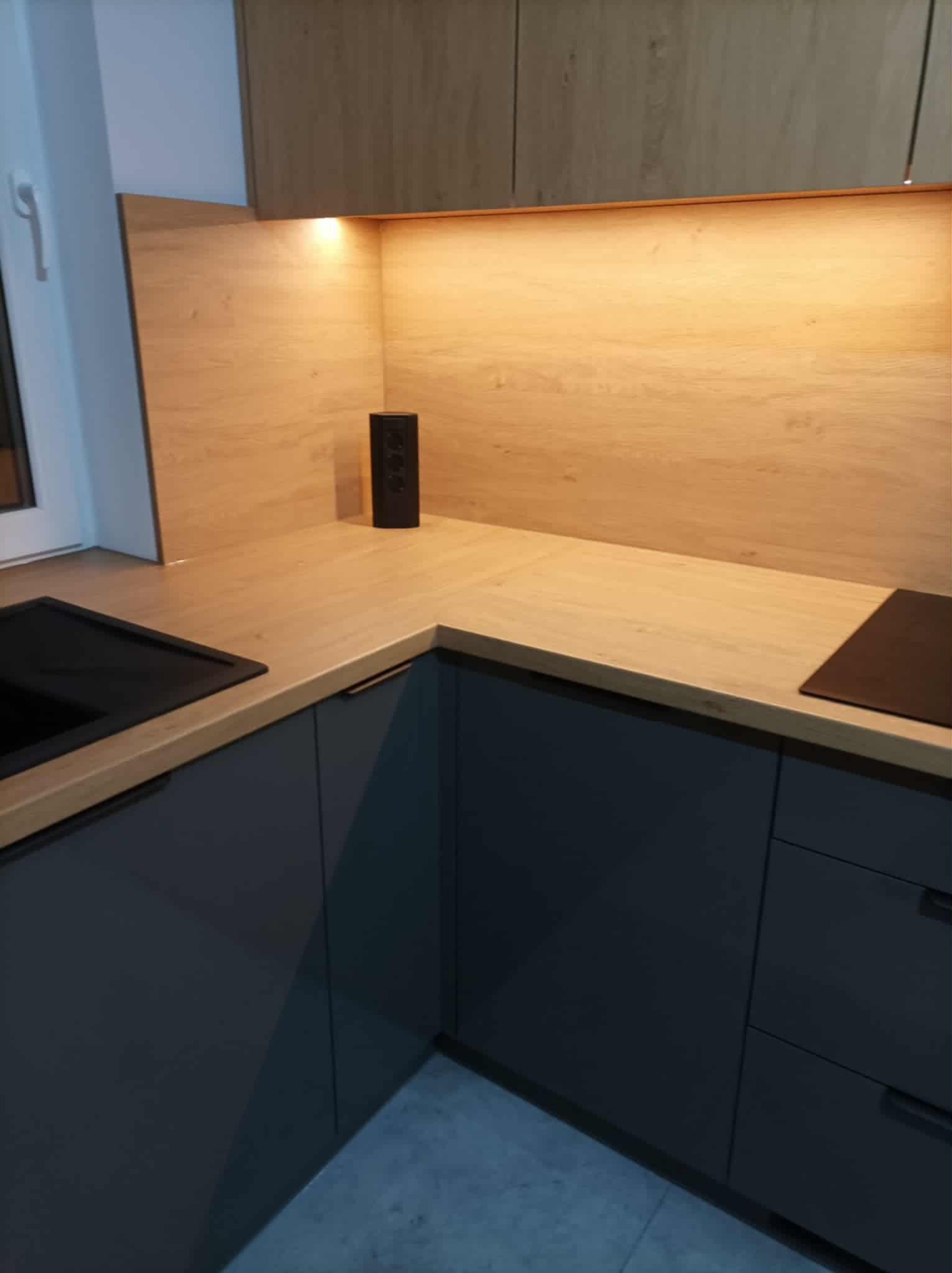 Nowoczesna kuchnia z szarymi szafkami i drewnianym blatem, oświetlona od spodu. Na blacie stoi czarny głośnik. Minimalistyczny design, widok z perspektywy narożnika.