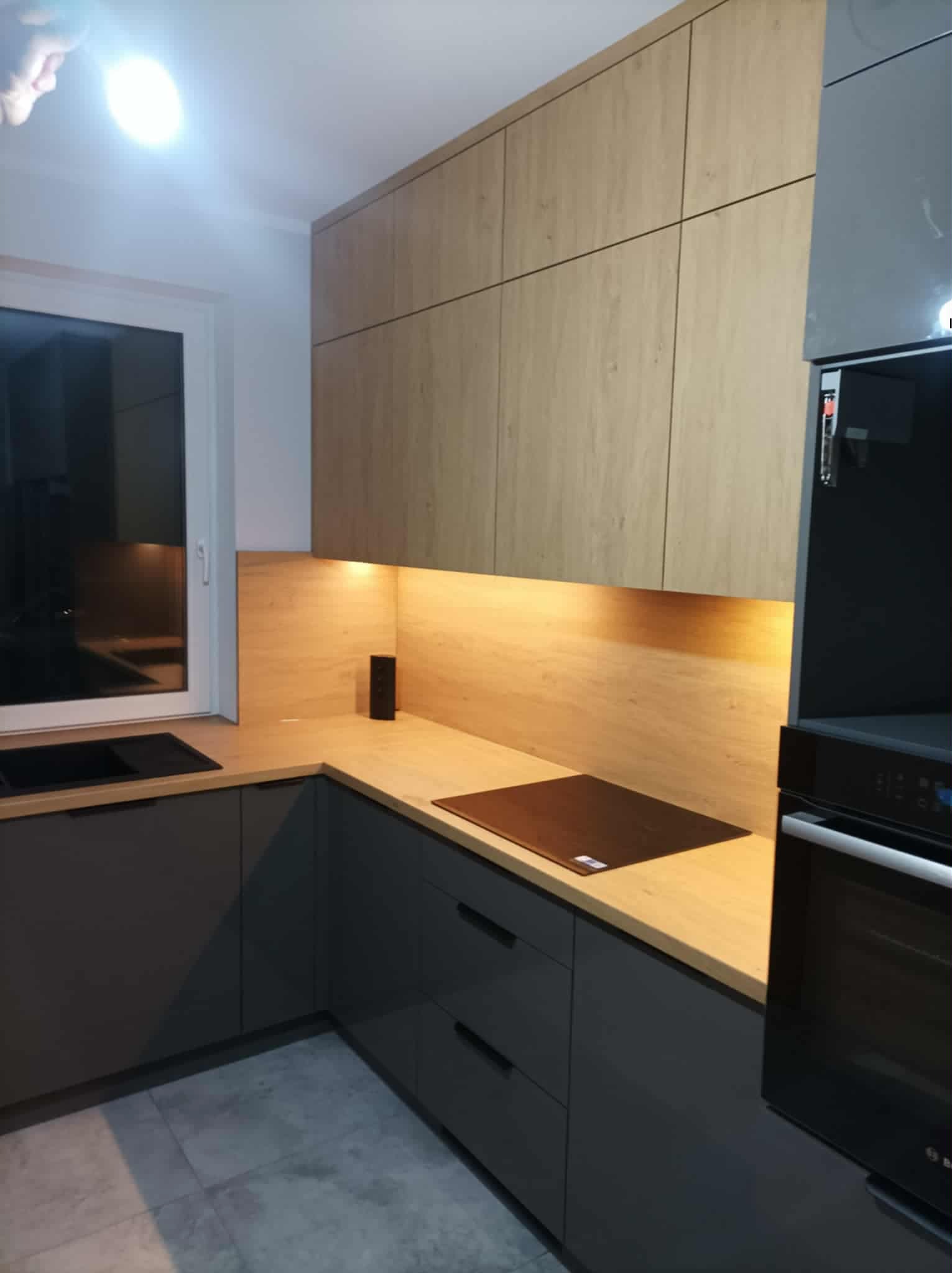 Nowoczesna kuchnia z szarymi szafkami dolnymi i drewnopodobnymi górnymi, podświetlona listwą LED. Widoczna płyta indukcyjna i piekarnik w zabudowie.