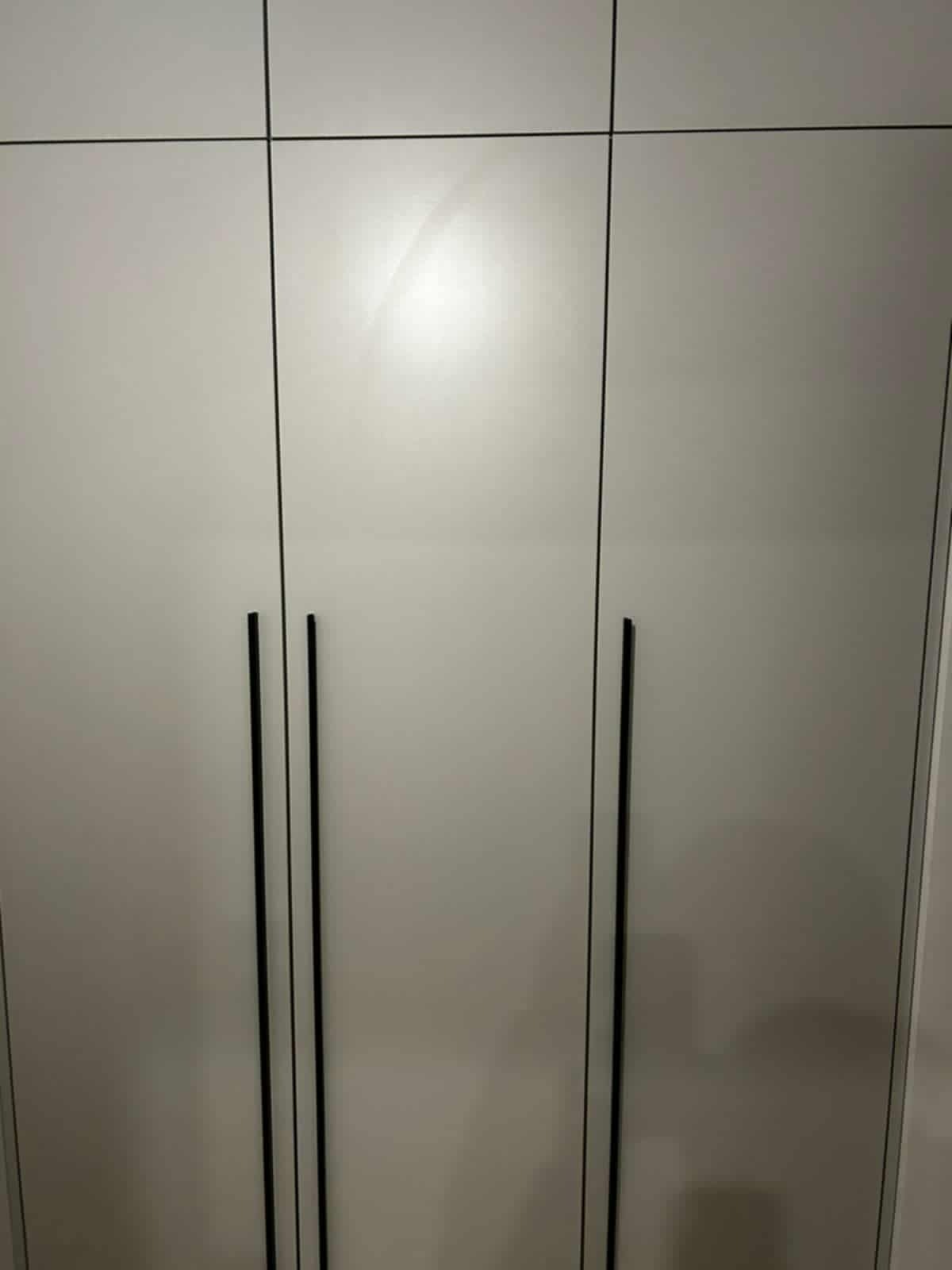 Minimalistyczna szafa wnękowa z trzema drzwiami w odcieniach szarości, z długimi, pionowymi, czarnymi uchwytami. Gładkie fronty, brak zdobień.